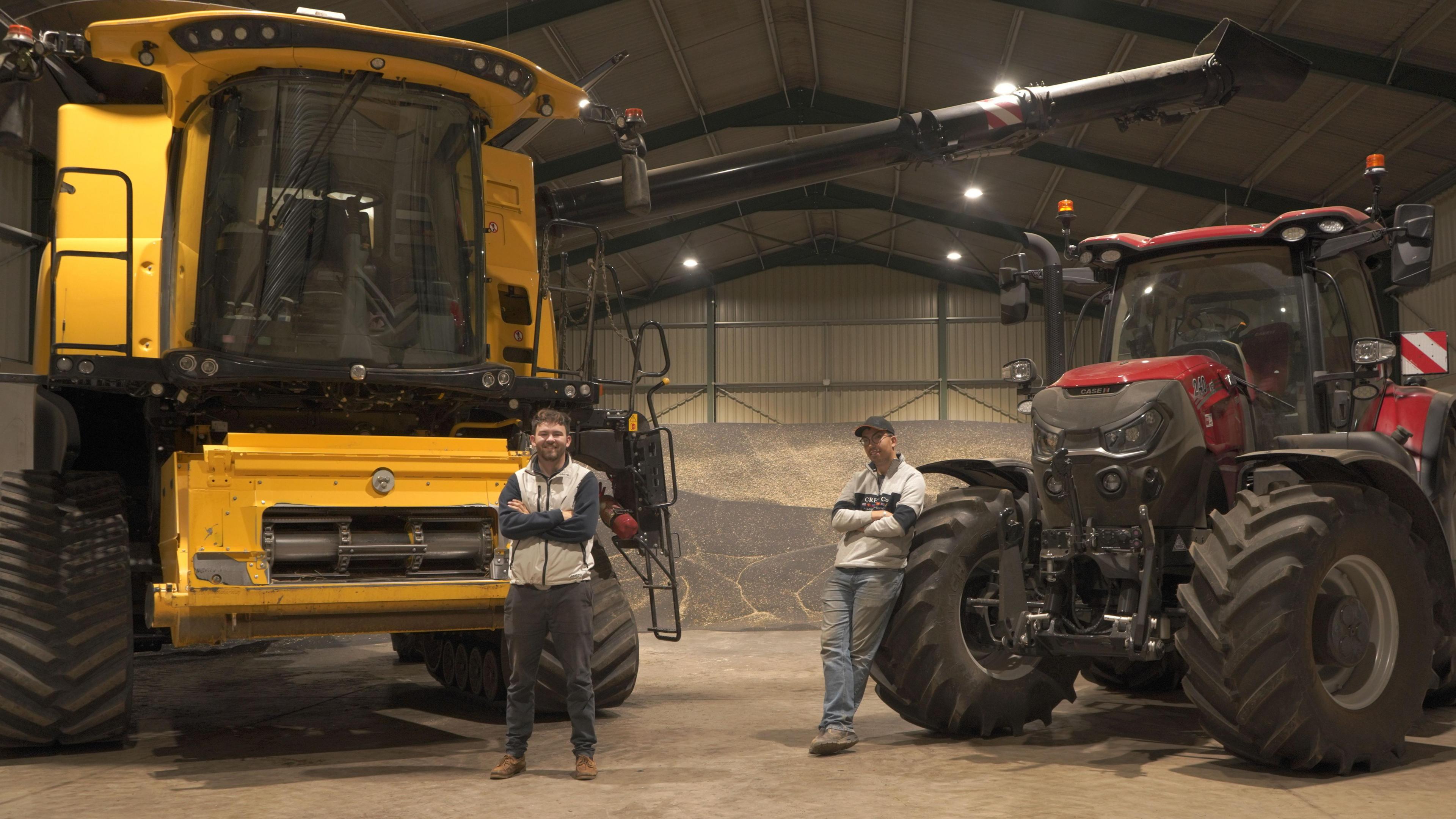 Two men stand with their arms folded in front of a yellow combine harvester and red tractor