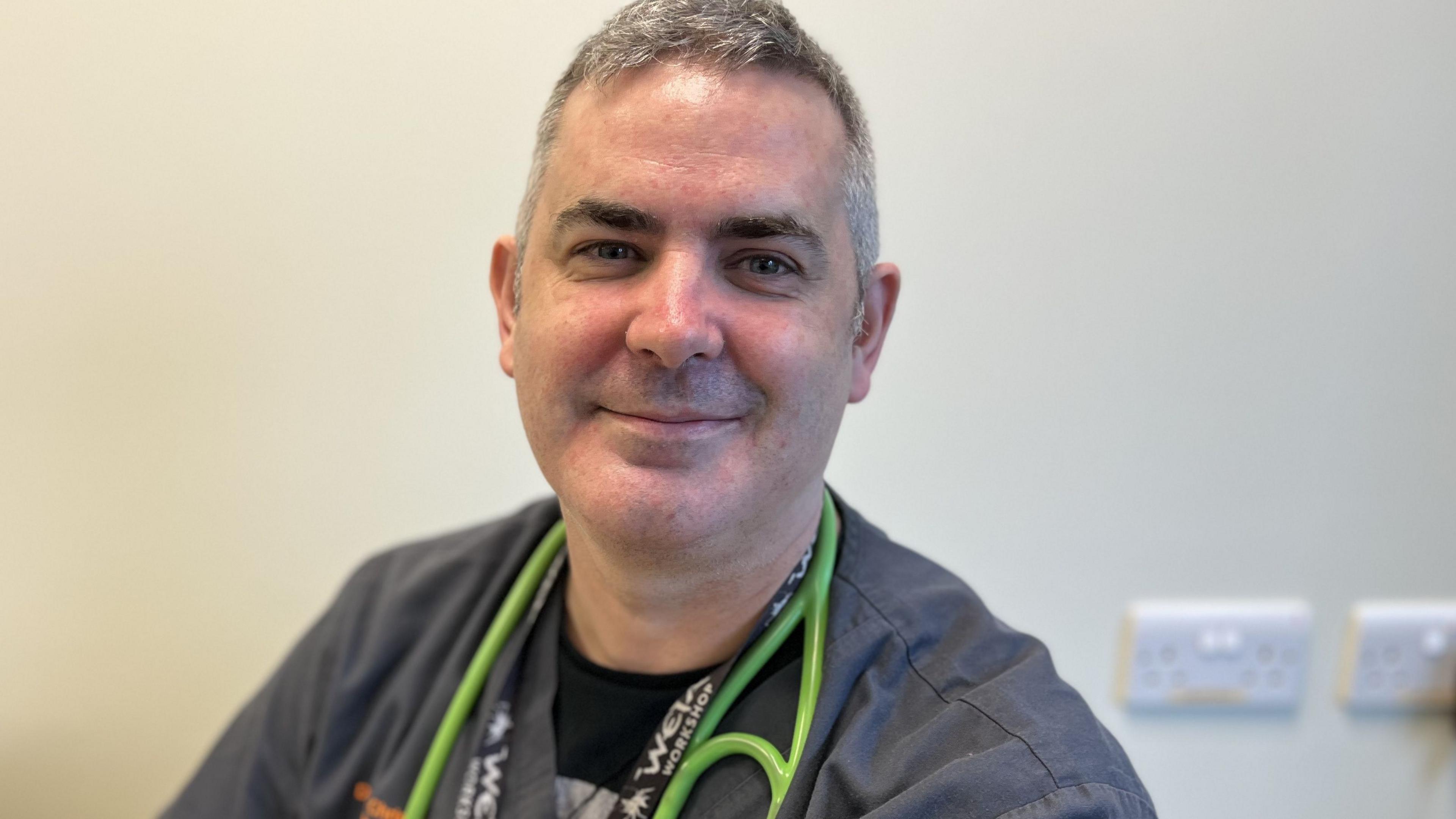 Doctor Chris Anderson, Paediatric Consultant on Sarum Ward smiling wearing grey scrubs with a green stethoscope around his neck