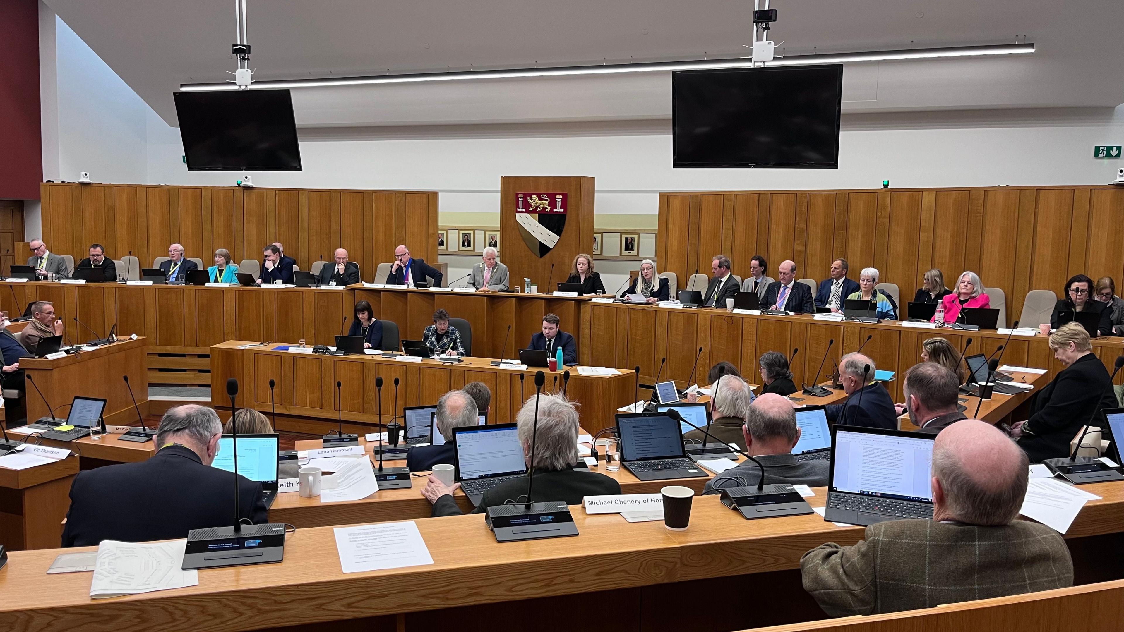 A curved council chamber with councillors sitting on rows of desks in front of the Norfolk county council logo. 