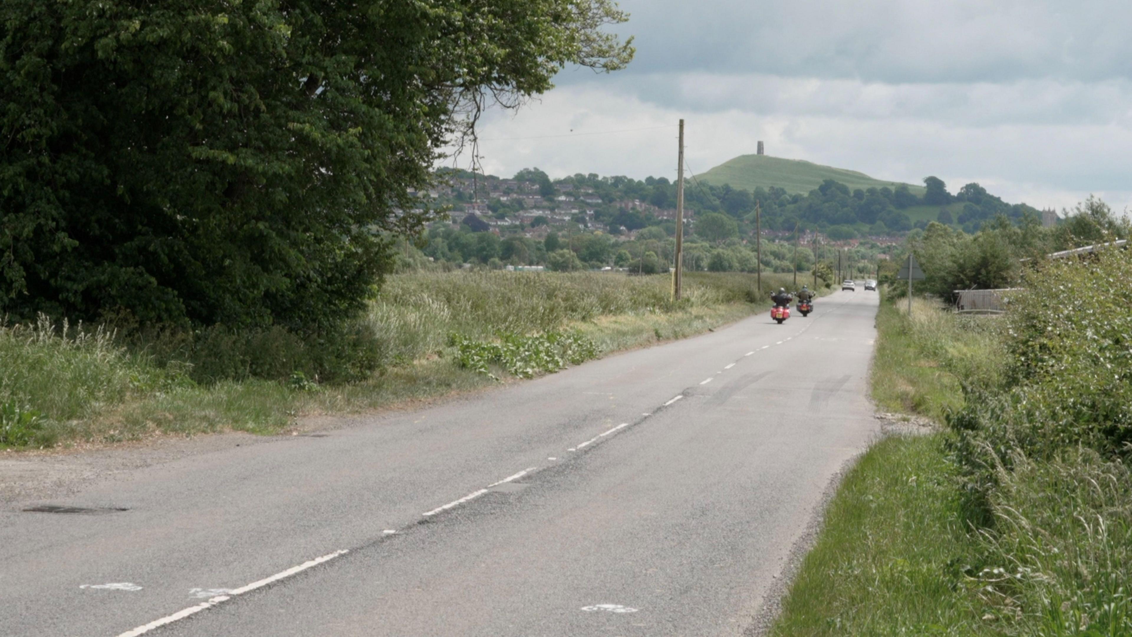 A straight country road with motorcyclists driving along and cars further away. Beyond the trees in the far distance is Glastonbury Tor.