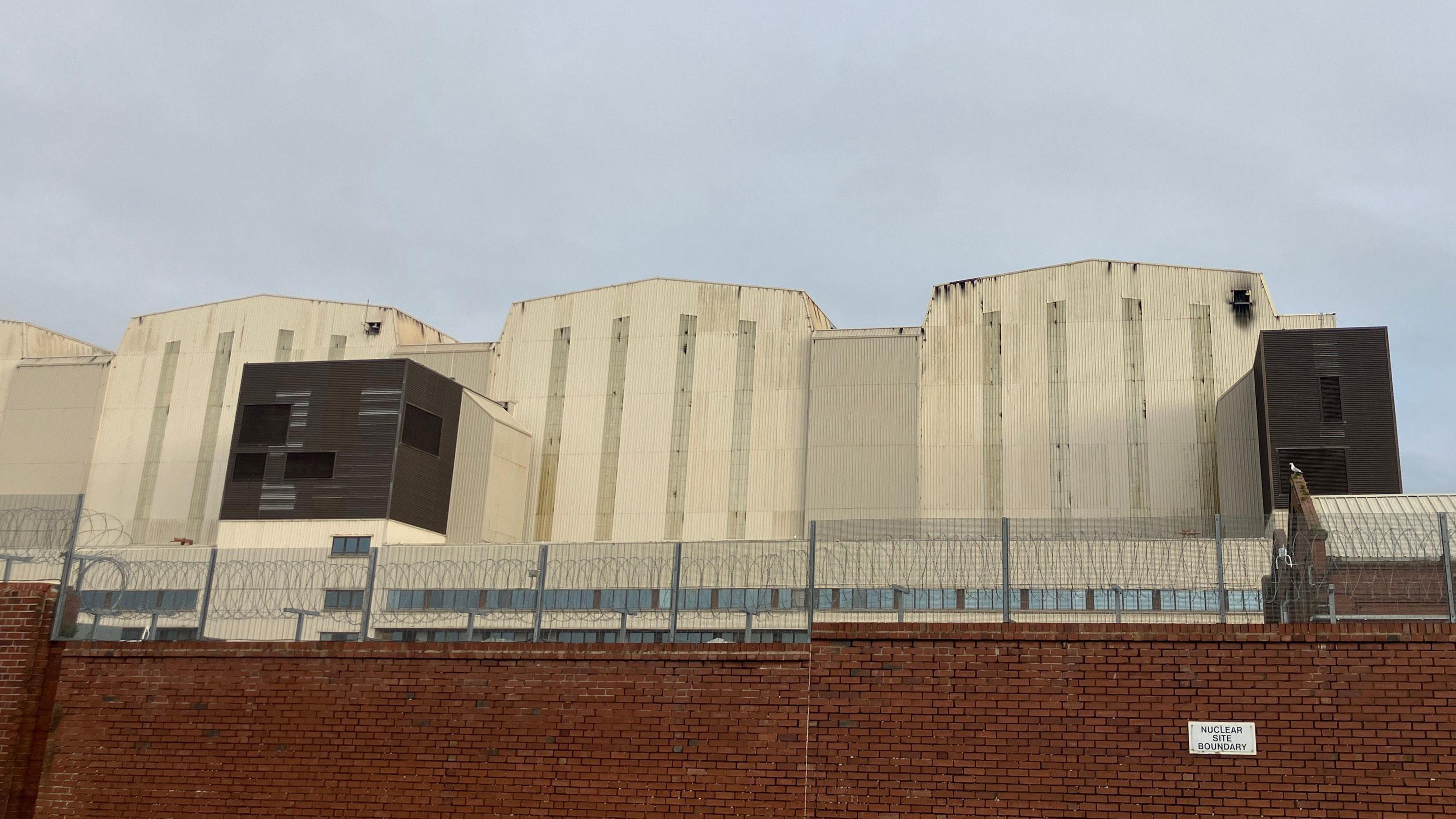 Photo of light grey bulky building with smoke damage to the top of the building. The skies are grey.