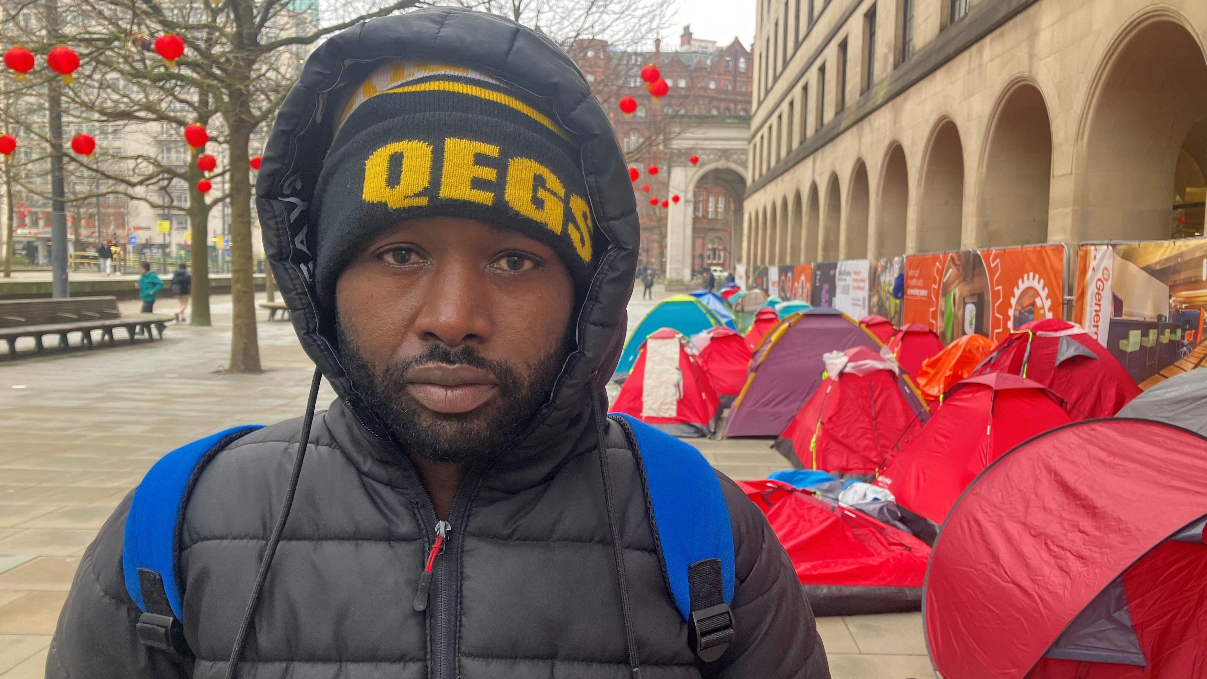 Adil Yusuf stands close to the red tents under a set of archways on St Peter's Squares in Manchester City Centre. Trees bearing red Chinese lanterns can be seen behind him. He is wearing a wooly hat, black puffer jacket and blue rucksack. 