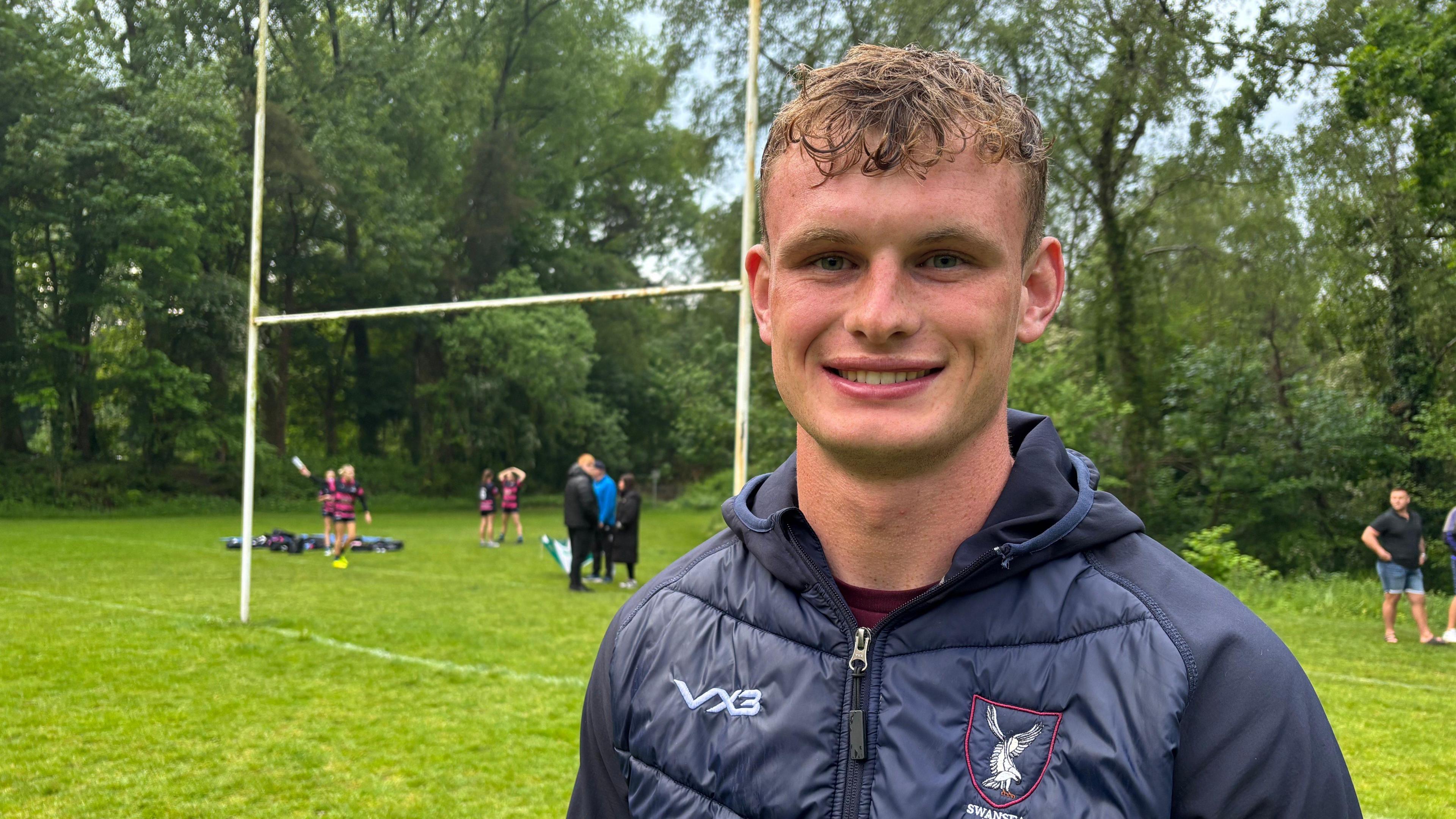 Joshua smiles at the camera. He has short, curly hair is wearing a navy down jacket with a club badge on the right hand side. There is a rugby pitch behind him where there are children and adults standing by the rugby goals.
