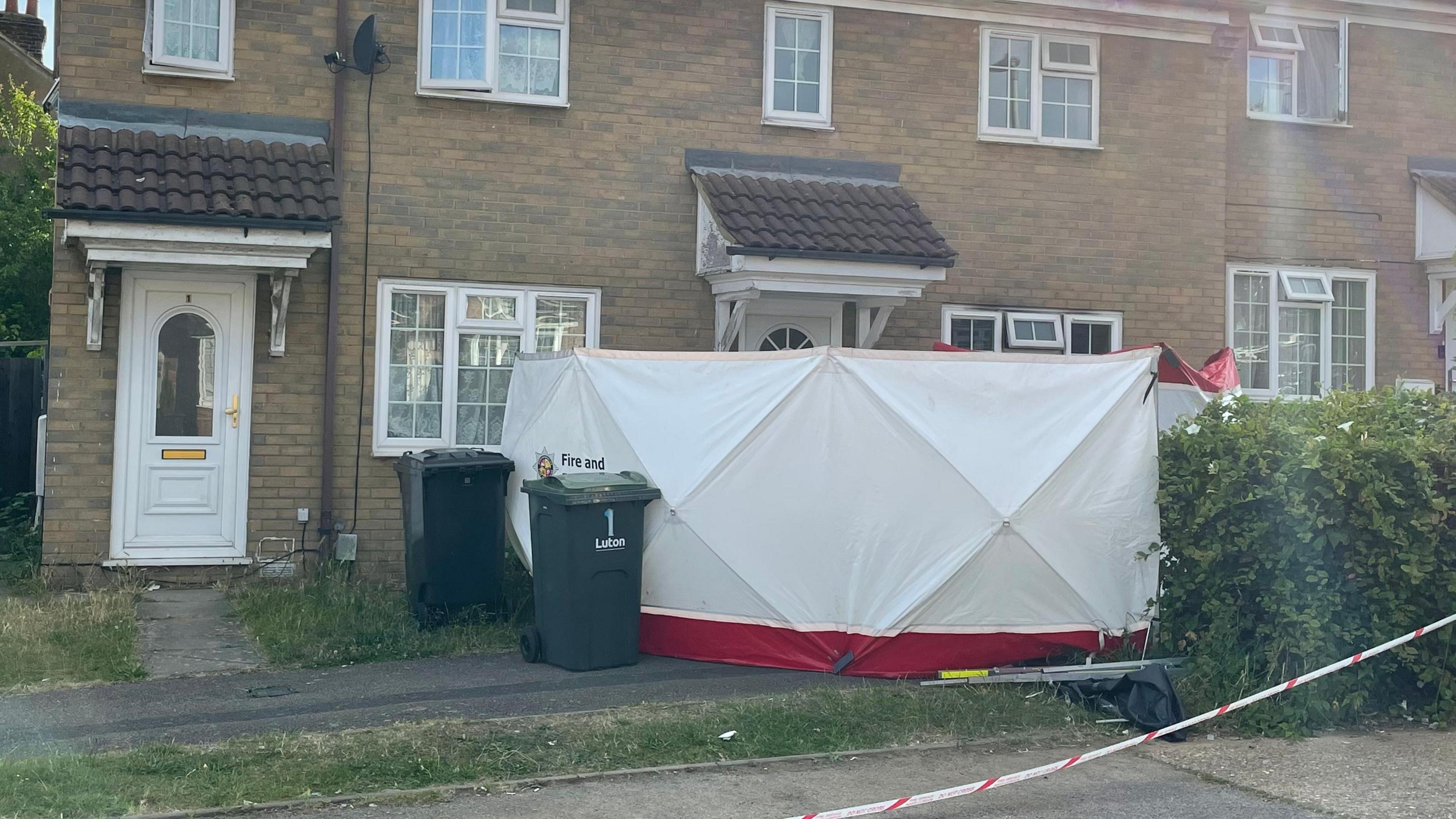 A white tent outside the entrance to the property, which also has two black wheelie bins out the front. Red and white tapes off the entrance to the house.