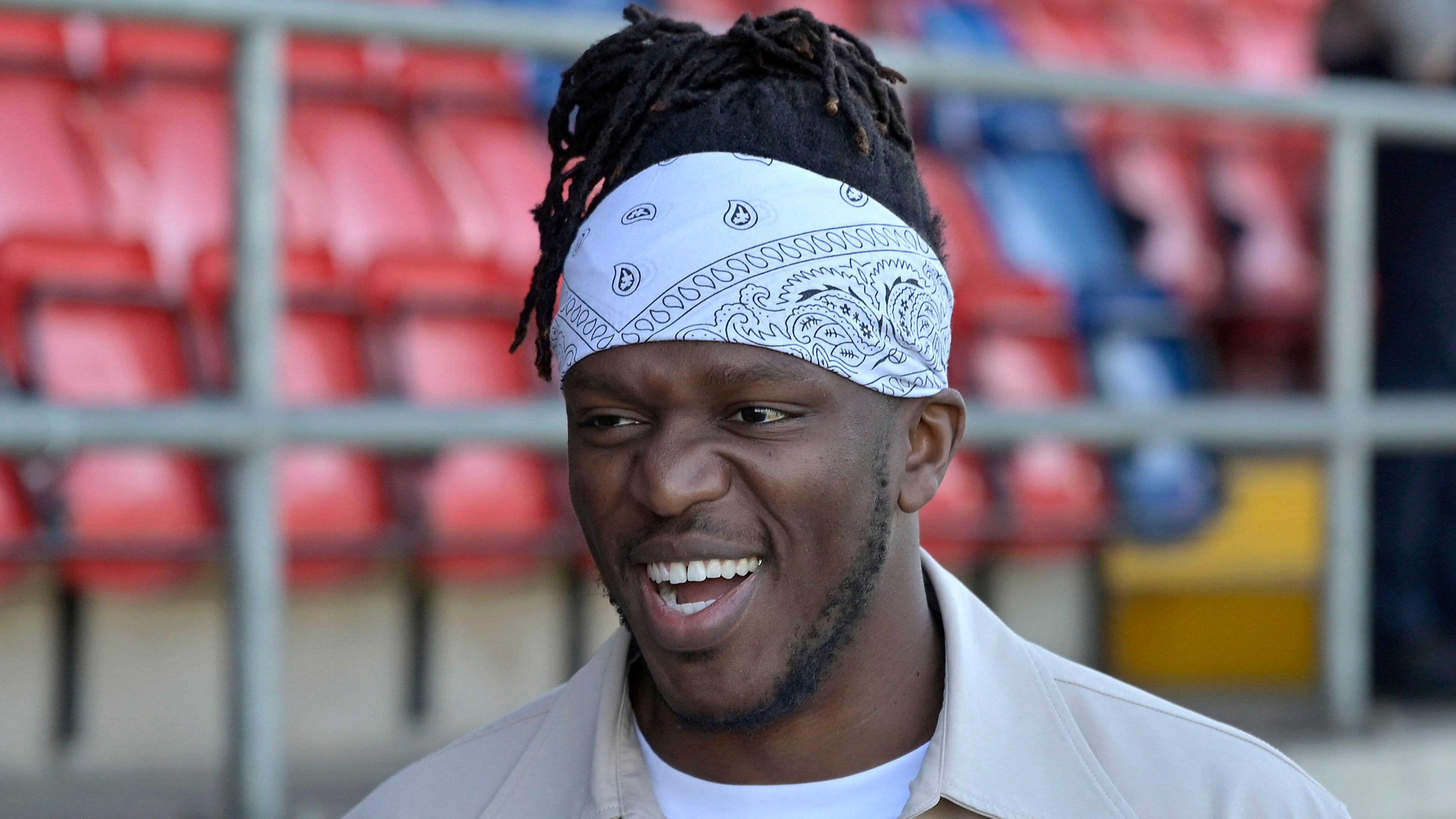 A smiling KSI wearing a beige jacket, white T-shirt and white bandana. The Dagenham & Redbridge stadium can be seen blurred behind him