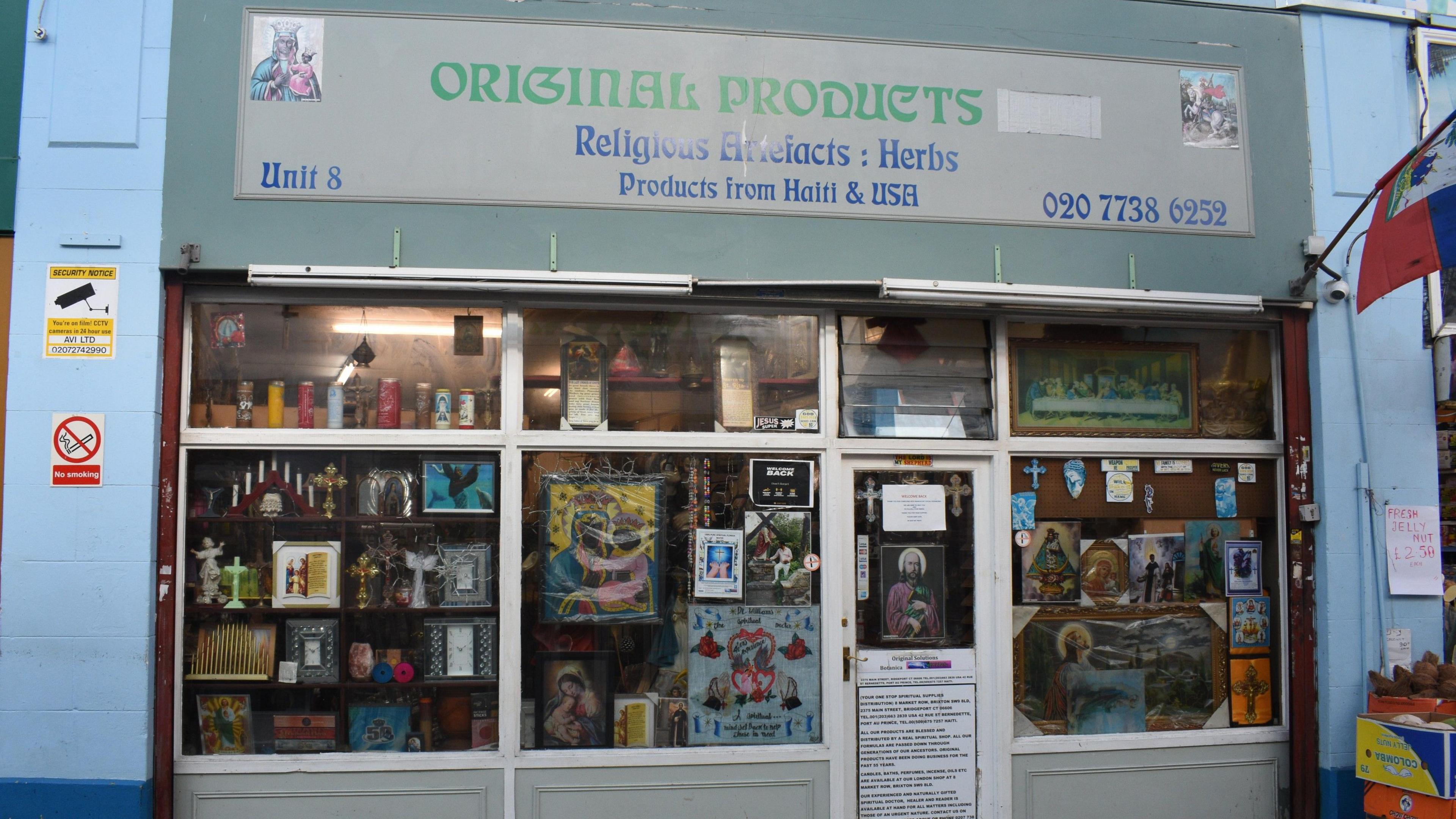 The facade of the Original Products store - with the wording Religious  Artefacts: Herbs and Products from Haiti and USA on the front. The windows and doors are full of paraphernalia. 