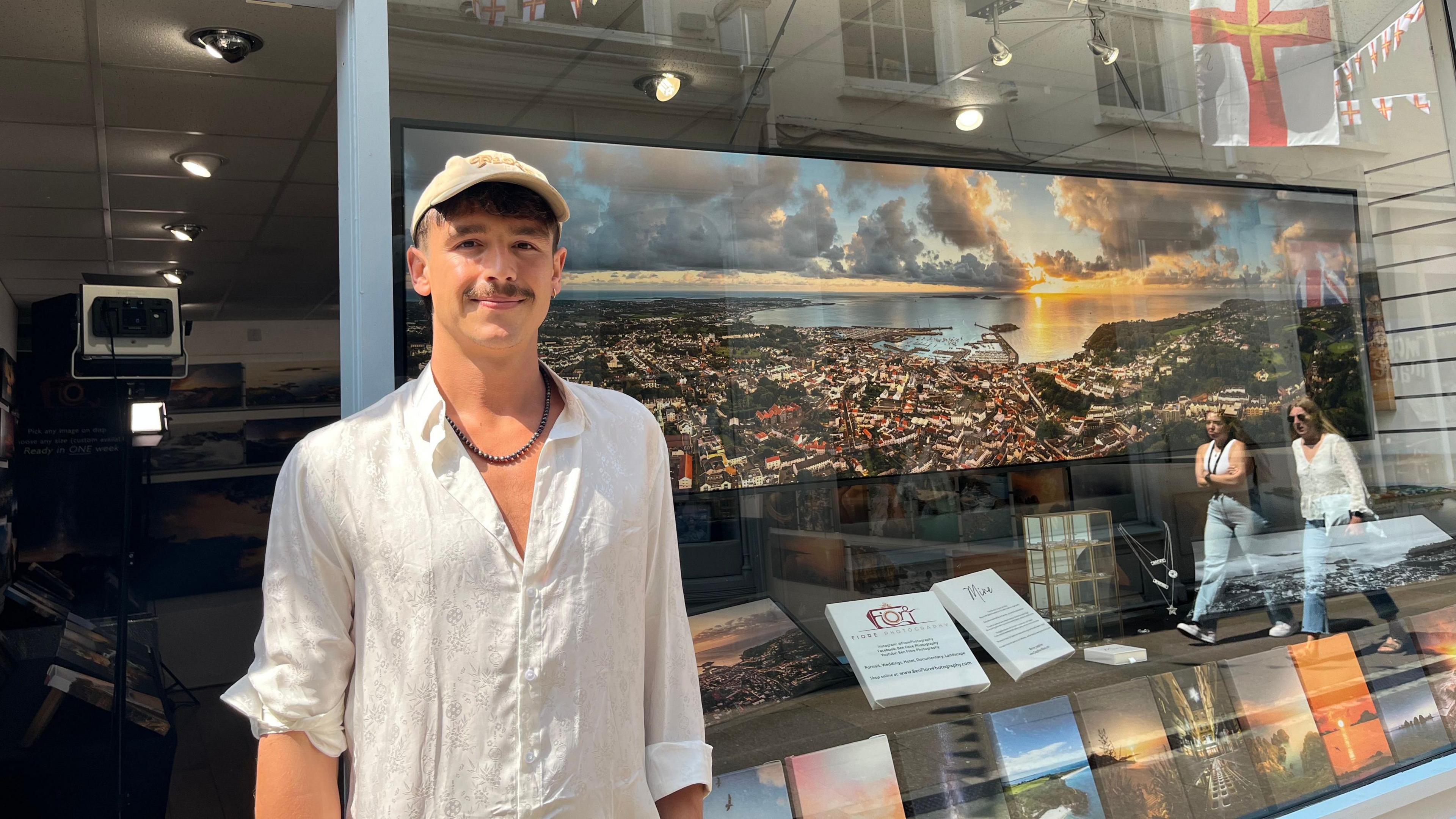 A man wearing a white shirt and a beige cap, smiling at the camera. He is standing beside a shop window with a panoramic photograph on display.