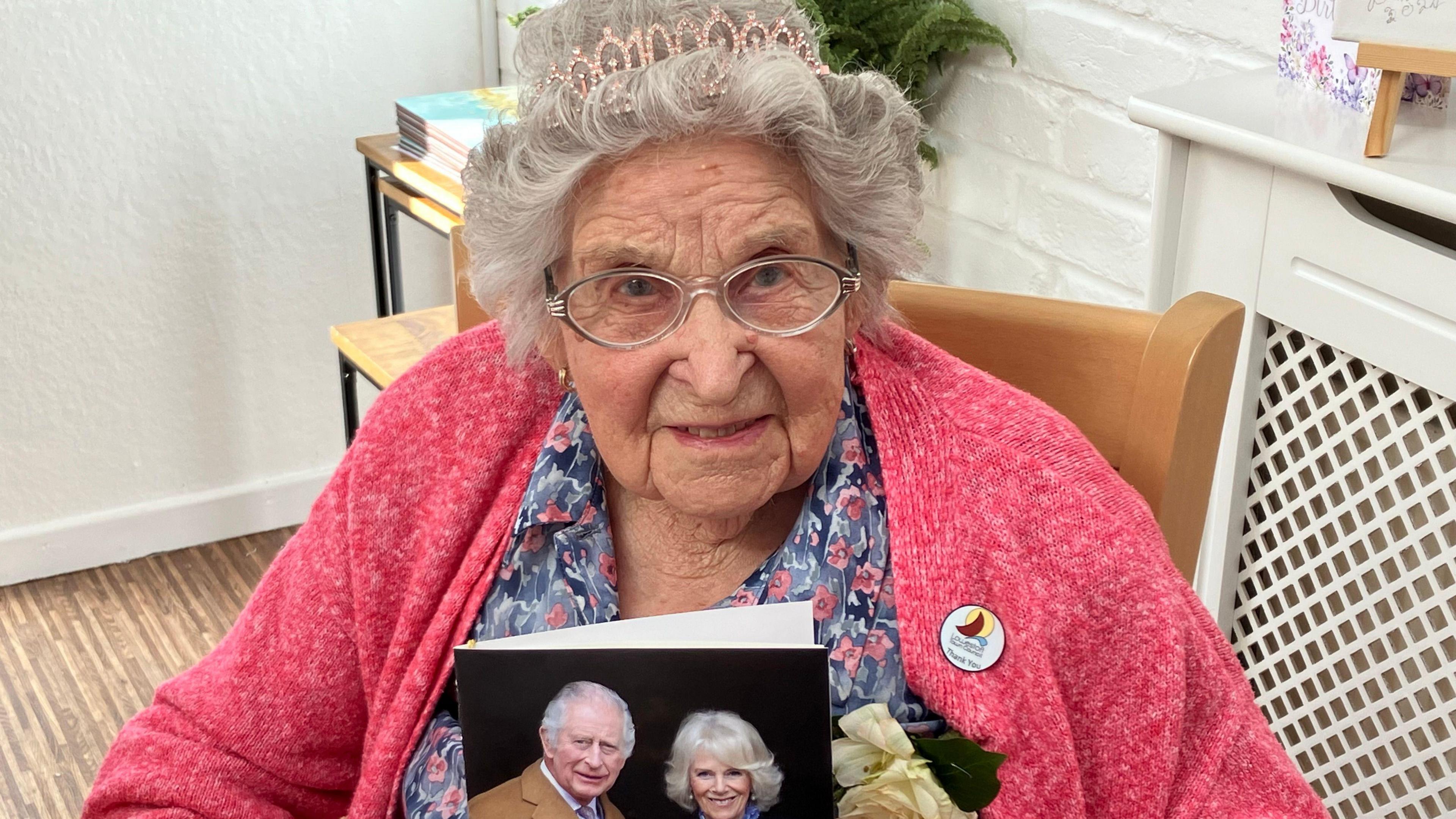 A woman wearing a tiara in her hair and a pink cardigan holds up a card from the King and Queen celebrating her 100th birthday