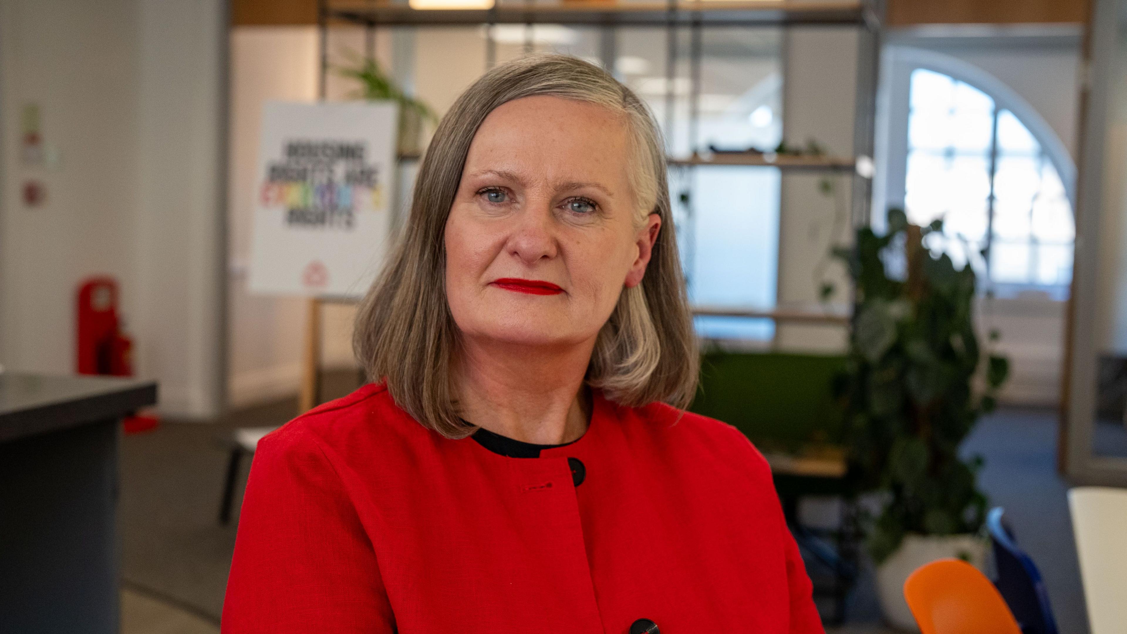 Alison Watson, a grey haired woman in a red jacket, is sitting in an office with desks, a pot plant and a window behind her.