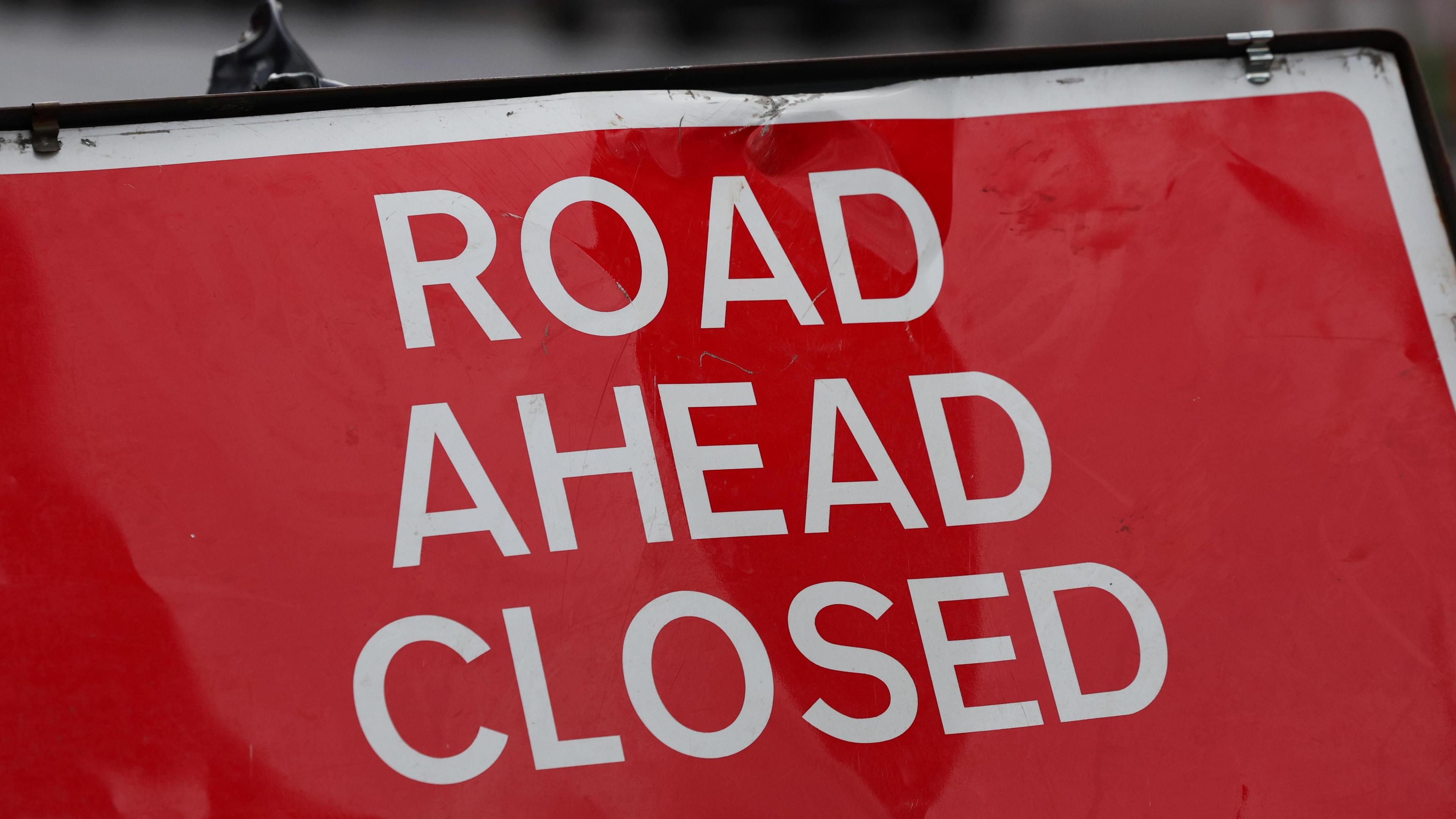 Close up of a 'road ahead closed' sign, with white writing on a red background. The sign is slightly battered.