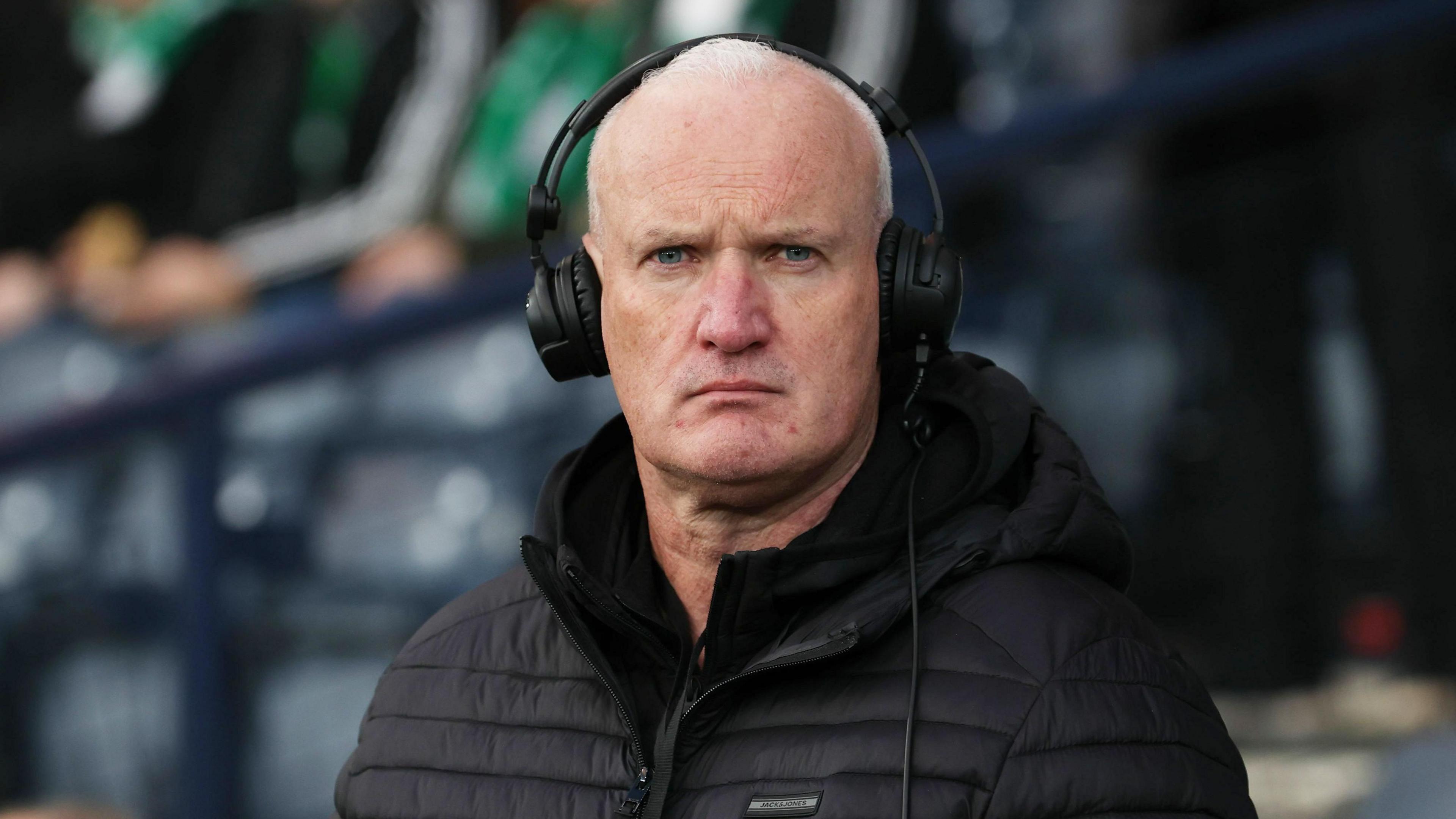 Kenny Macintyre in a football stadium wearing a black puffer jacket and black headphones