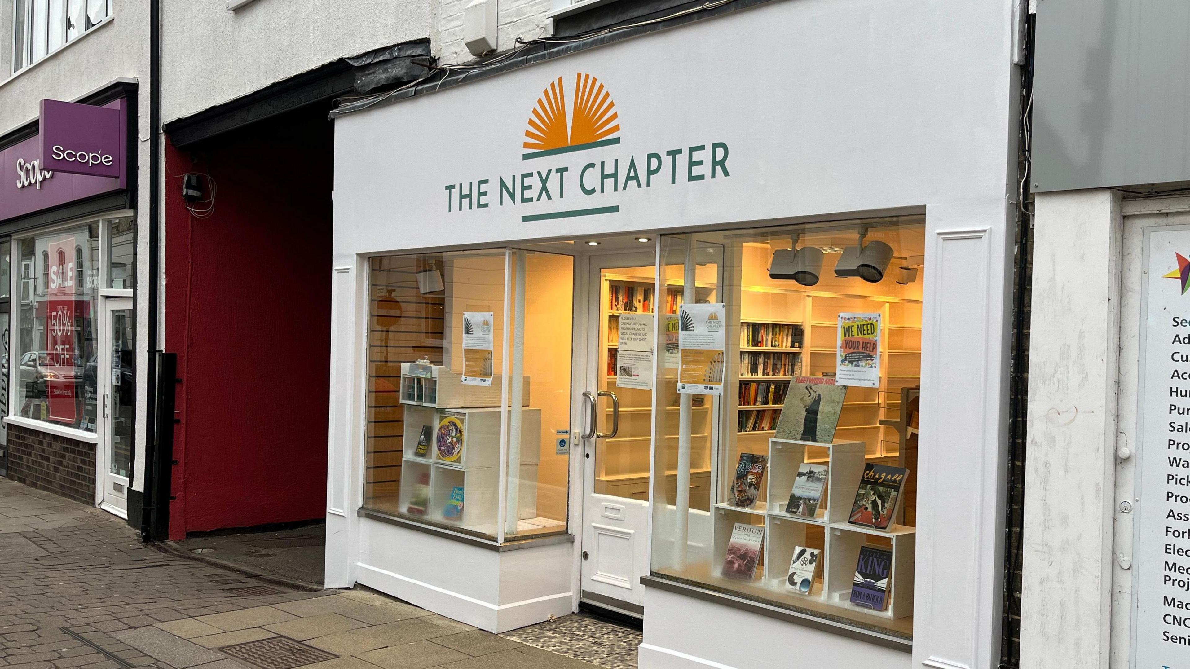 The shop is seen just off centre to the right of the picture. The building is white, and has glass windows either side of a door in the middle. A green sign above the door reads 'The Next Chapter'.