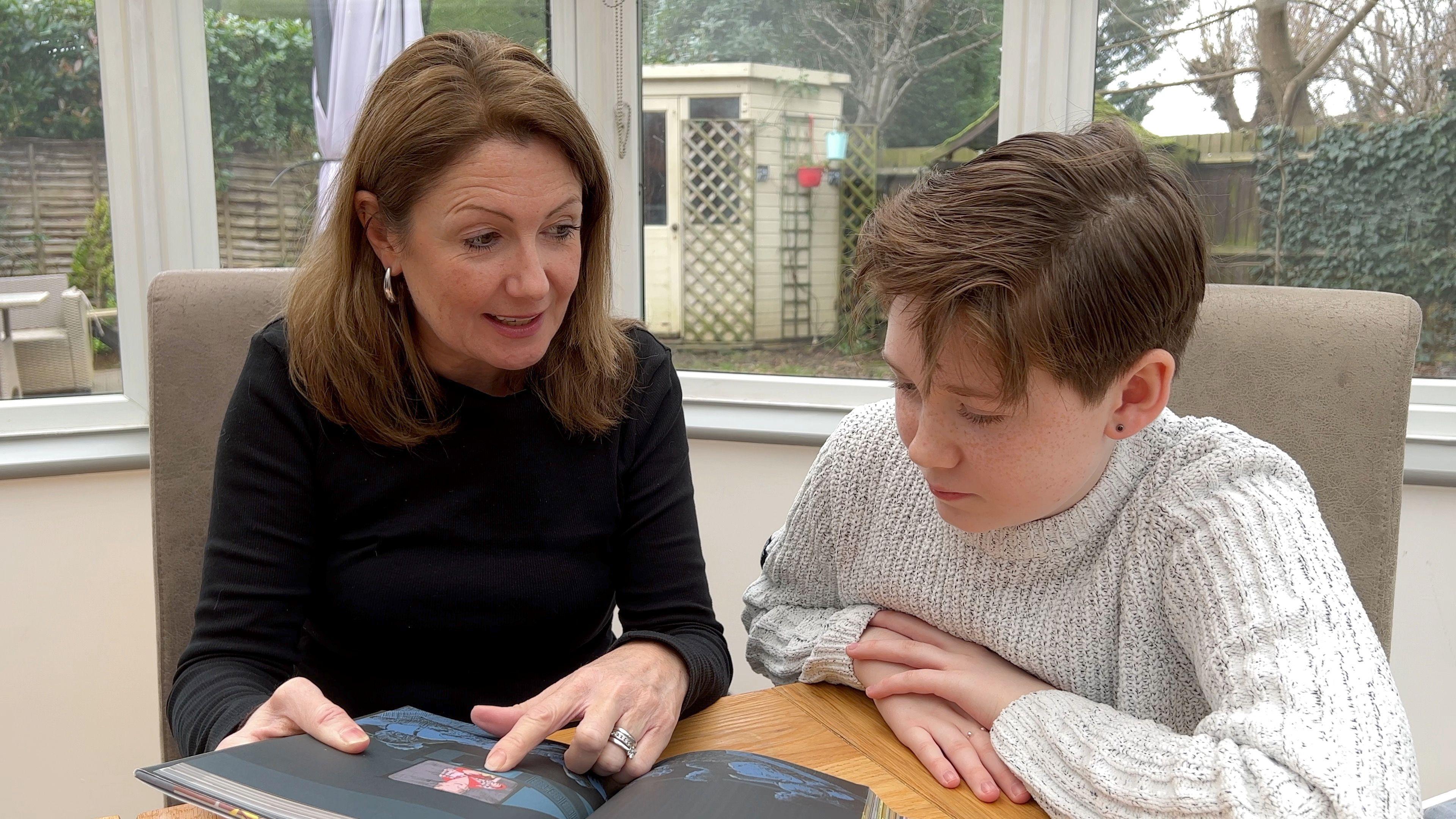 (L-R) Leigh is smiling and wearing a black roll neck top and Albie is wearing a white knitted jumper both are sitting at a table and looking at a book.