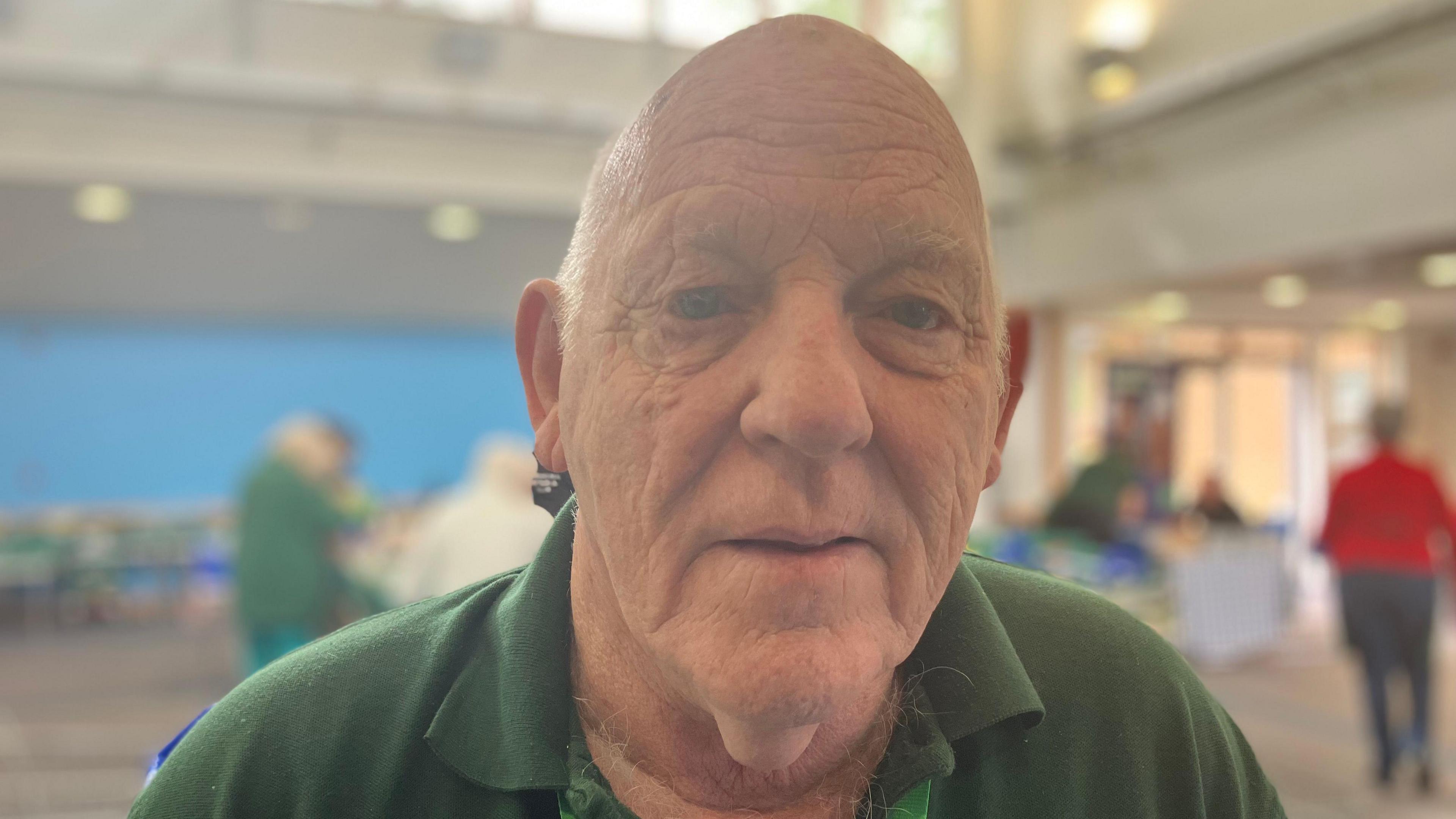 A head-and-shoulders photo of a bald man wearing a green polo shirt. In soft focus behind him there's a church hall with people milling around.