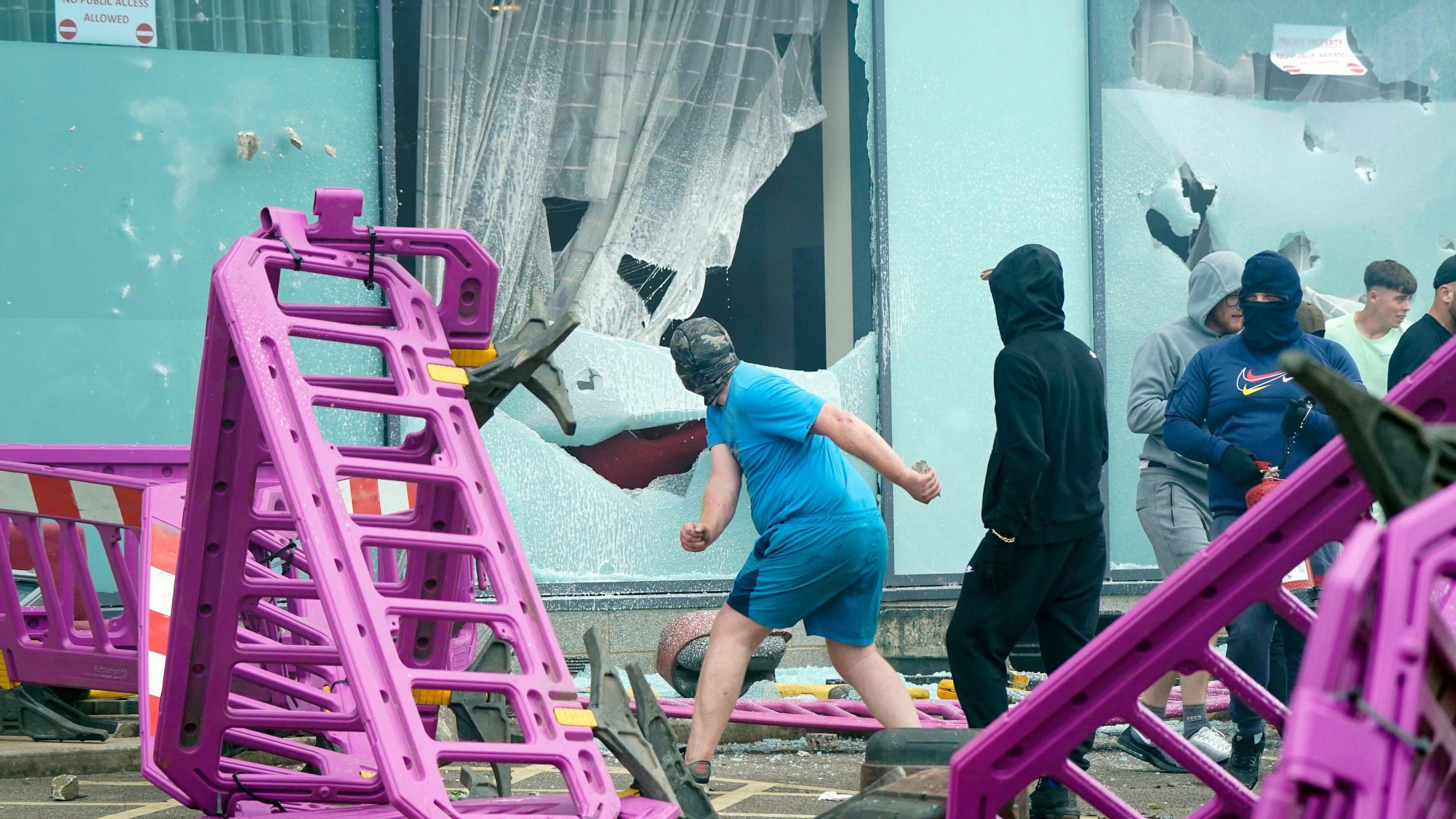 A man in a blue t-shirt throws an item through a hotel window to smash it. Purple barriers are on the floor around him.