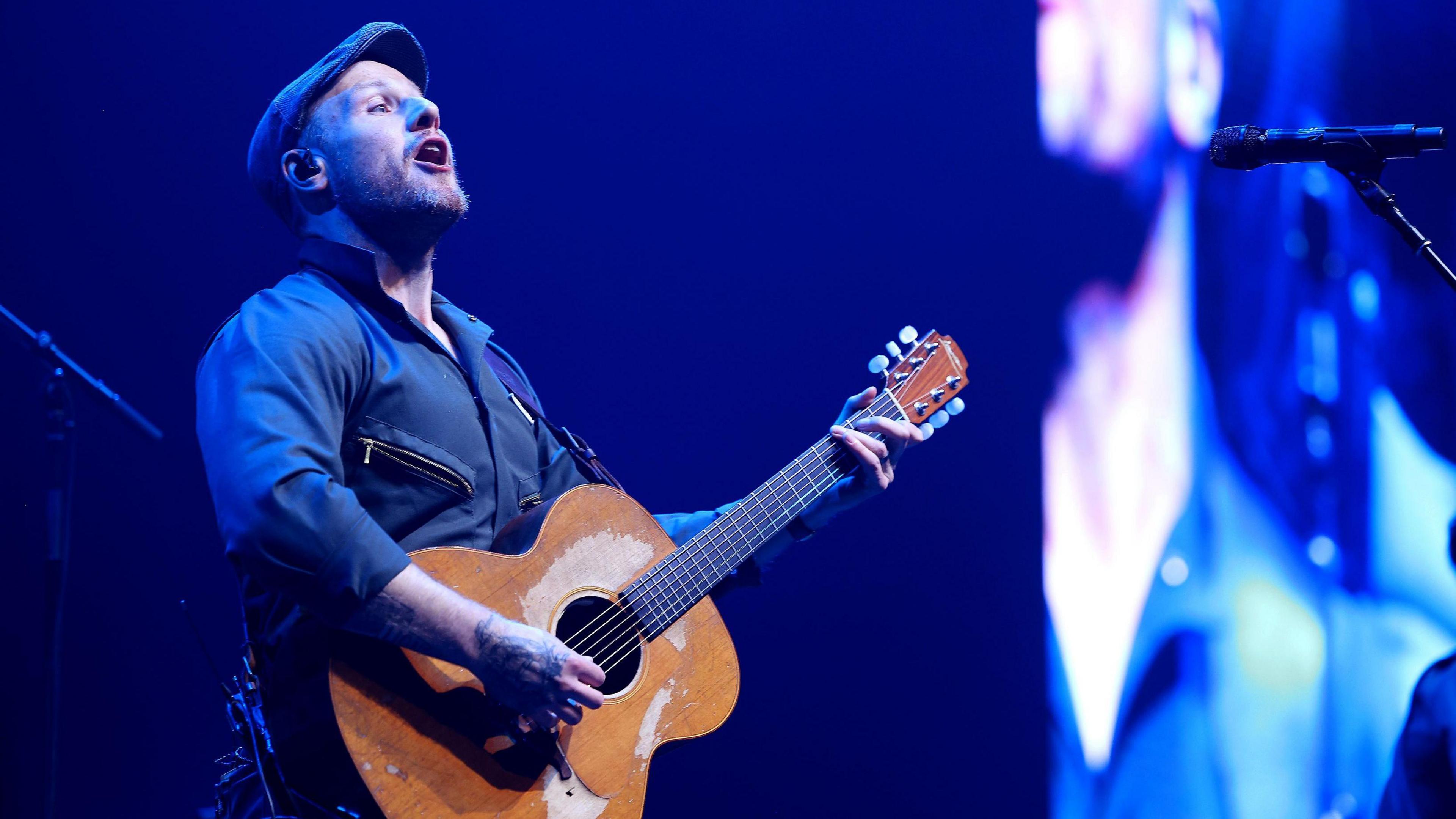 Chris Llewellyn is singing out to the crowd while he is on-stage. He is holding a guitar strapped over his shoulder and is wearing a navy shirt and waistcoat with zips on the left and right breast pockets. He has a light beard and is wearing a navy flat cap.