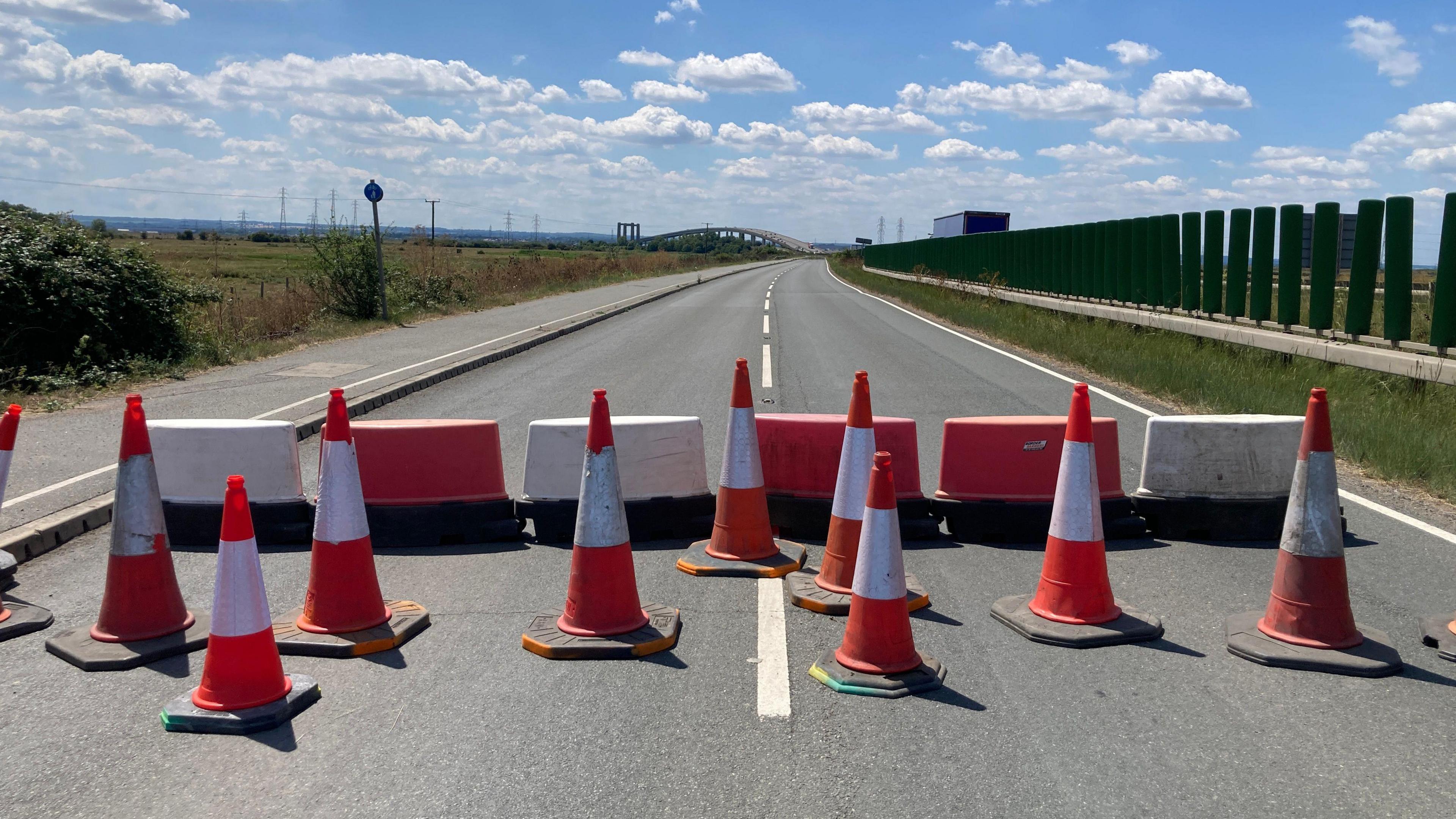 Ten red and white traffic cones and five red and white moveable barriers block access to a dual carriageway on a sunny day.
