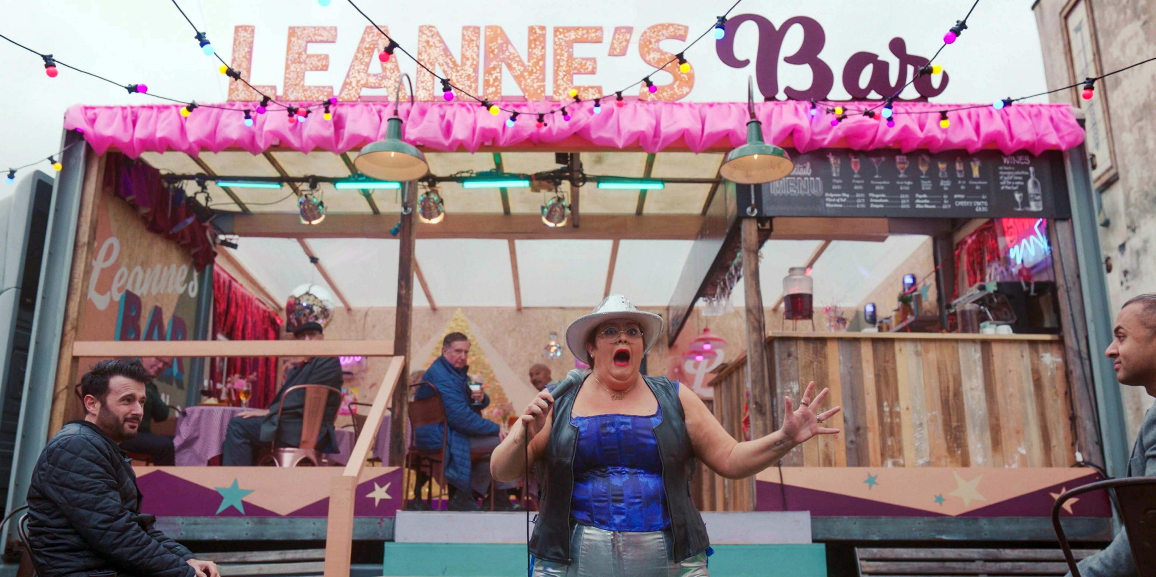 Leanne (Jayde Adams) is singing, wearing a blue corset style top, with a gilet, silver jeans and a white cowgirl hat