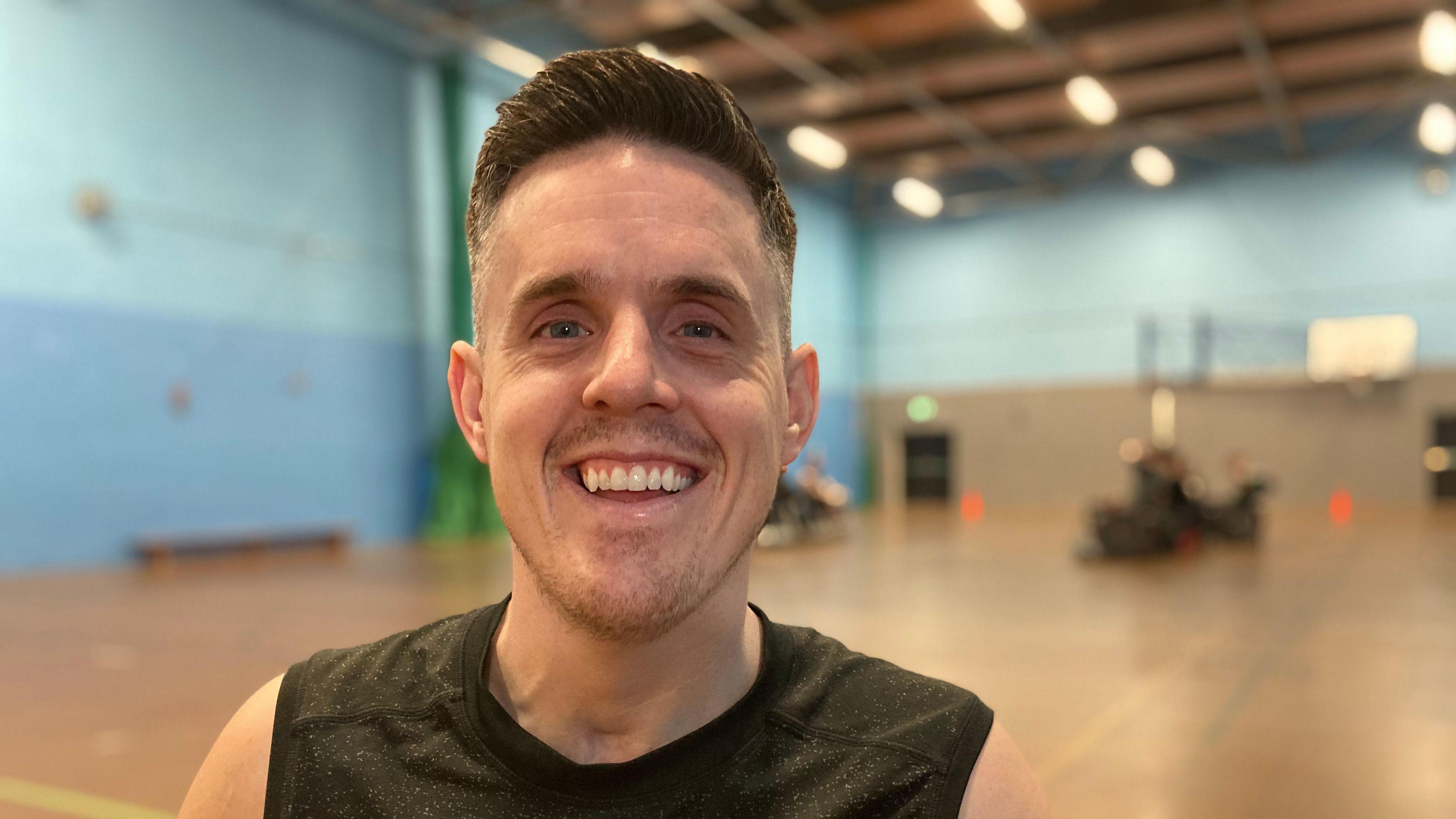A man sat in a sports hall is smiling at the camera, he is wearing a grey vest.