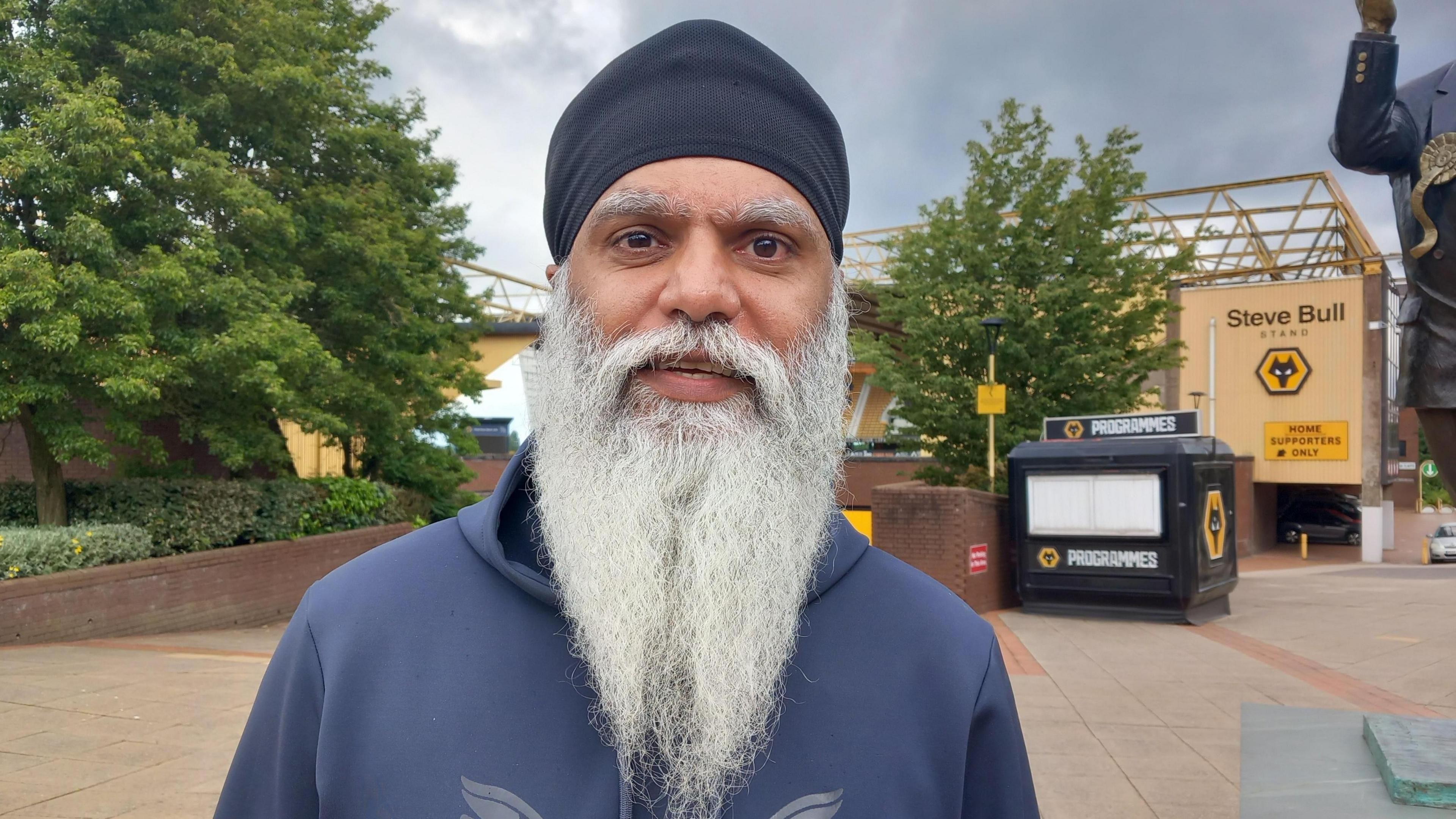 Manny Singh Kang smiling in front of Molineux Stadium wearing a blue hoodie 