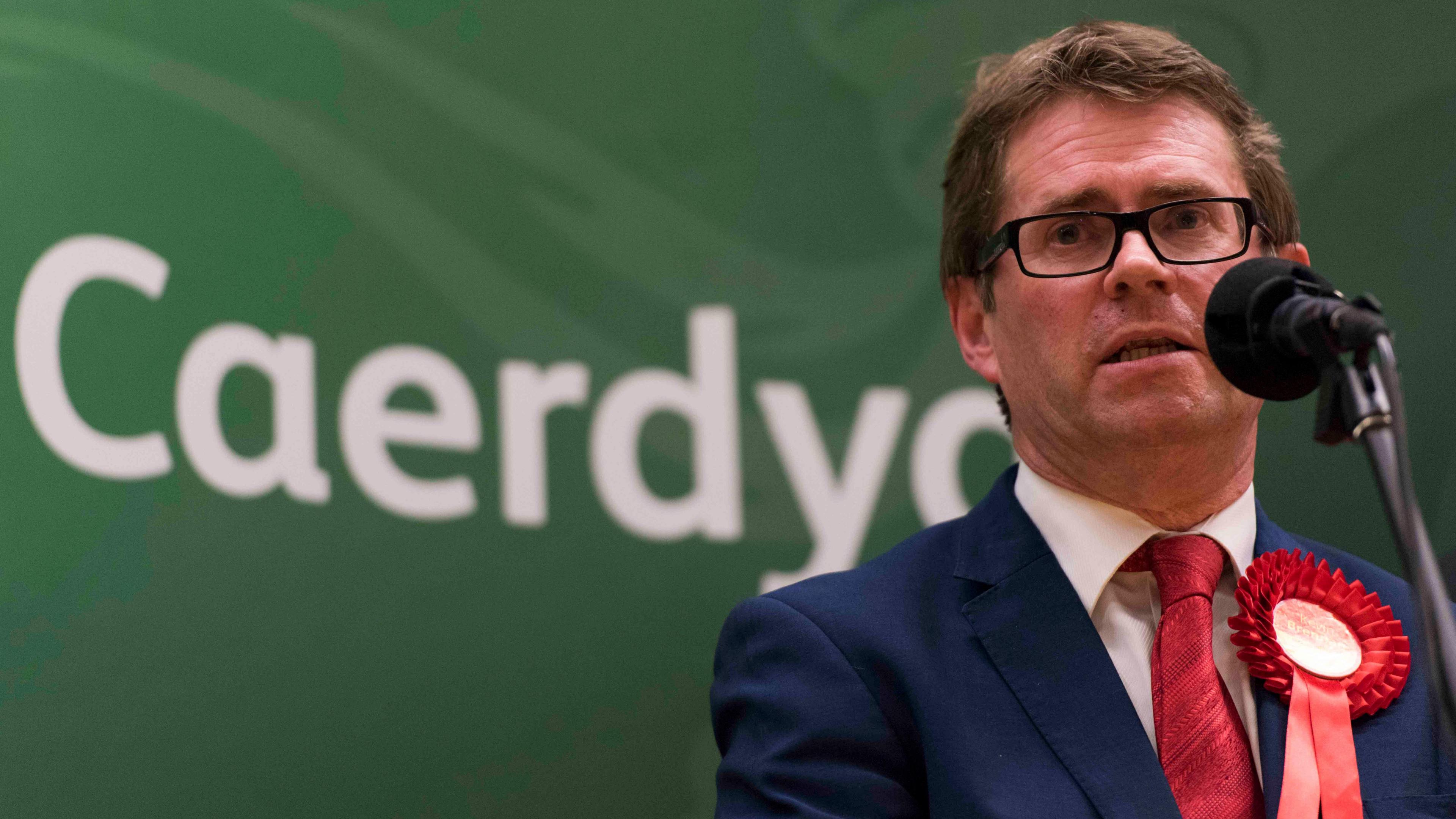 Kevin Brennan standing at a microphone with a red floret and Cardiff on a sign behind him in Welsh 