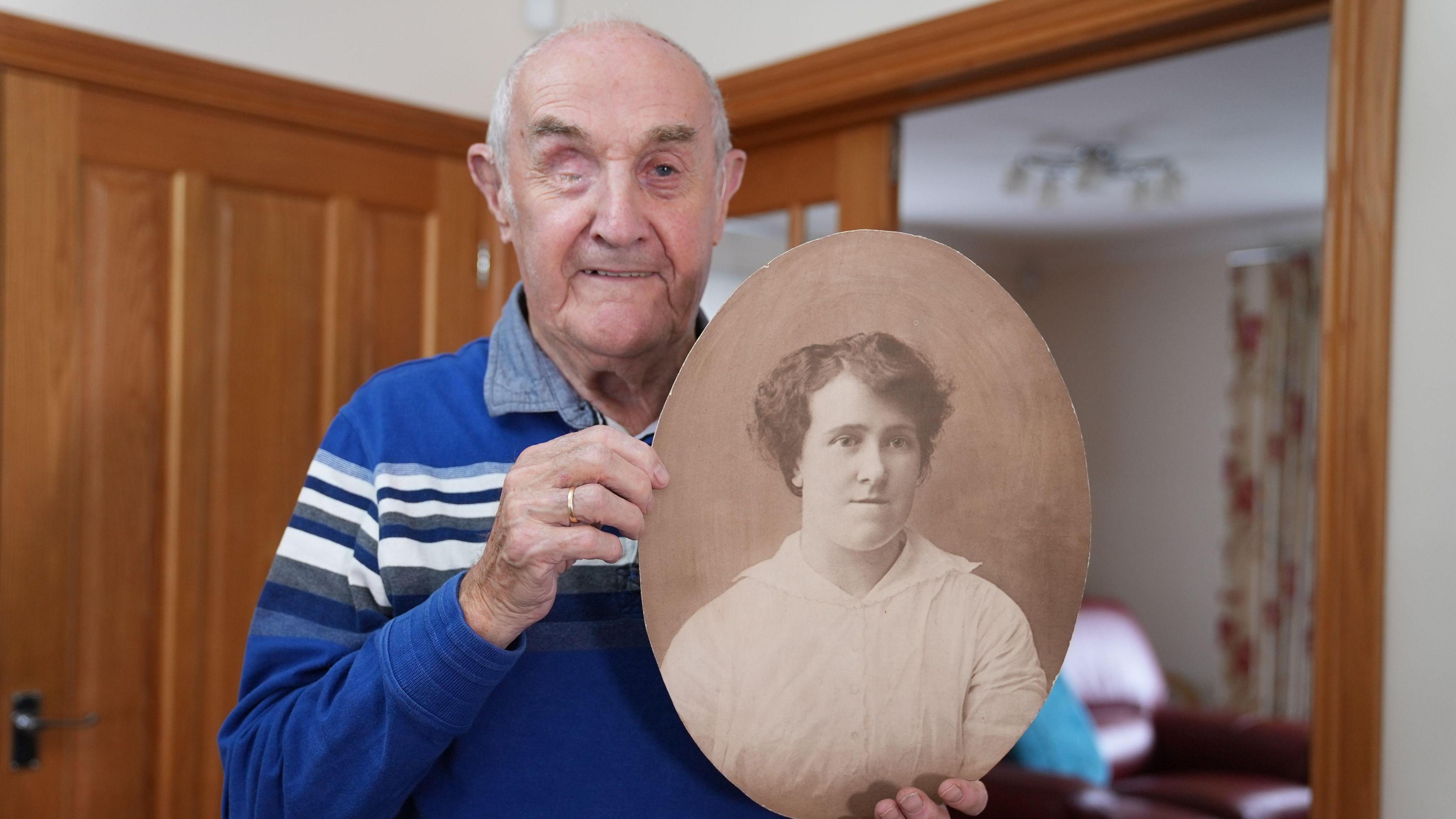 Mae ŵyr Mabel, Eddie Wulff, yn gwenu wrth ddal llun o'i famgu. Mae e'n gwisgo crys glas wrth gael ei lun wedi tynu yn ei gartref yng Nghasnewydd. Mae llun ei fam-gu yn sepia.