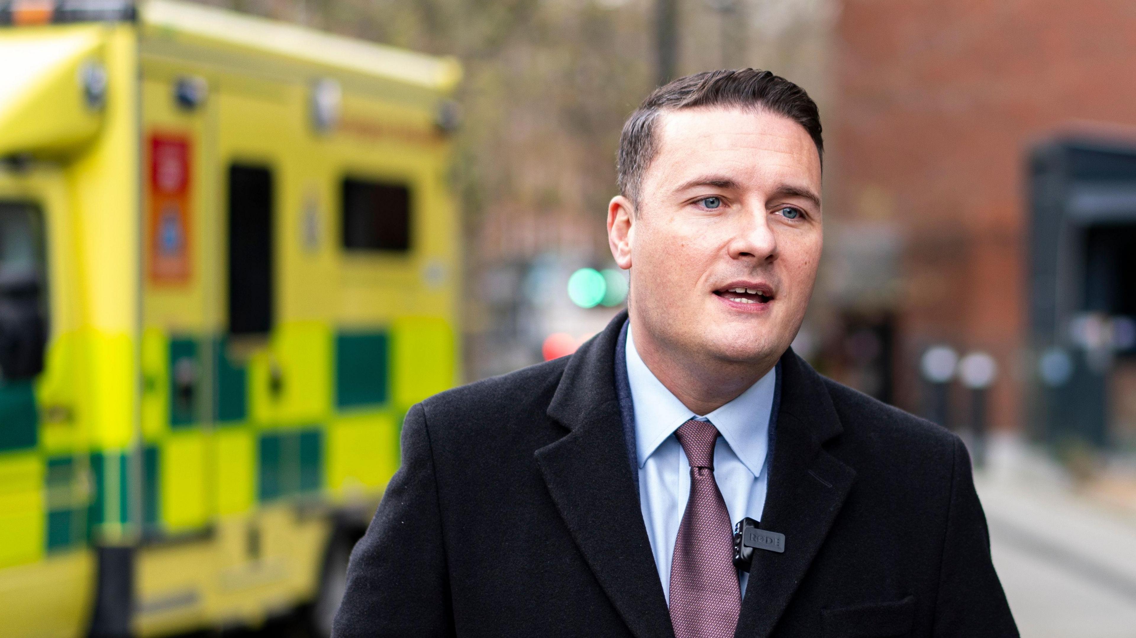 Health Secretary Wes Streeting wearing a shirt, tie and overcoat and standing in front of an ambulance.