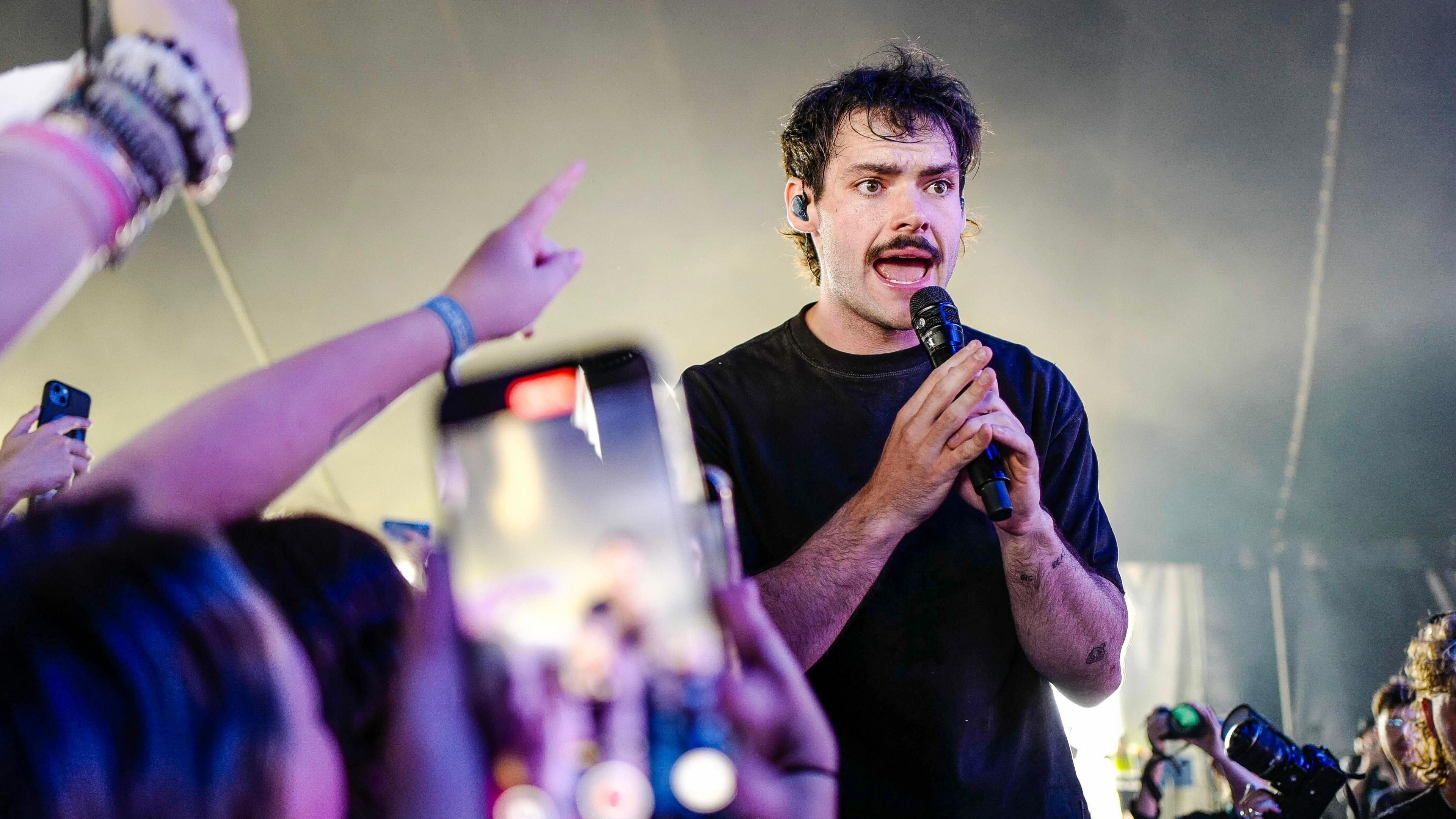 James Marriott, a young man with dark, ruffled hair and a moustache, holds both hands around a mic, wearing an an expression of concentration. Hands and phones held up by the crowd are seen near him.