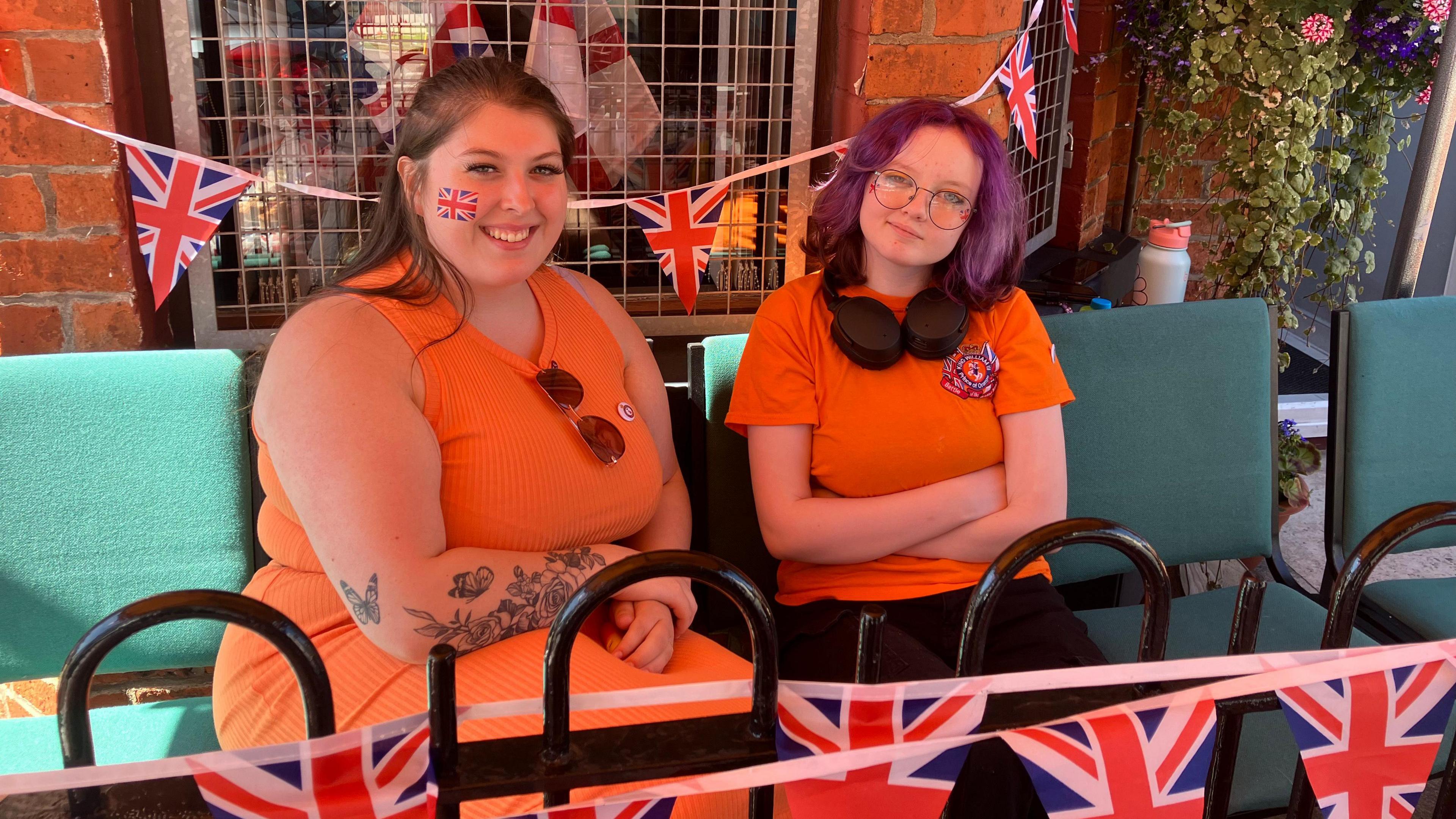 Two women are sitting outside on chairs. They are both wearing orange outfits and have Union Jack temporary tattoos on their faces. 