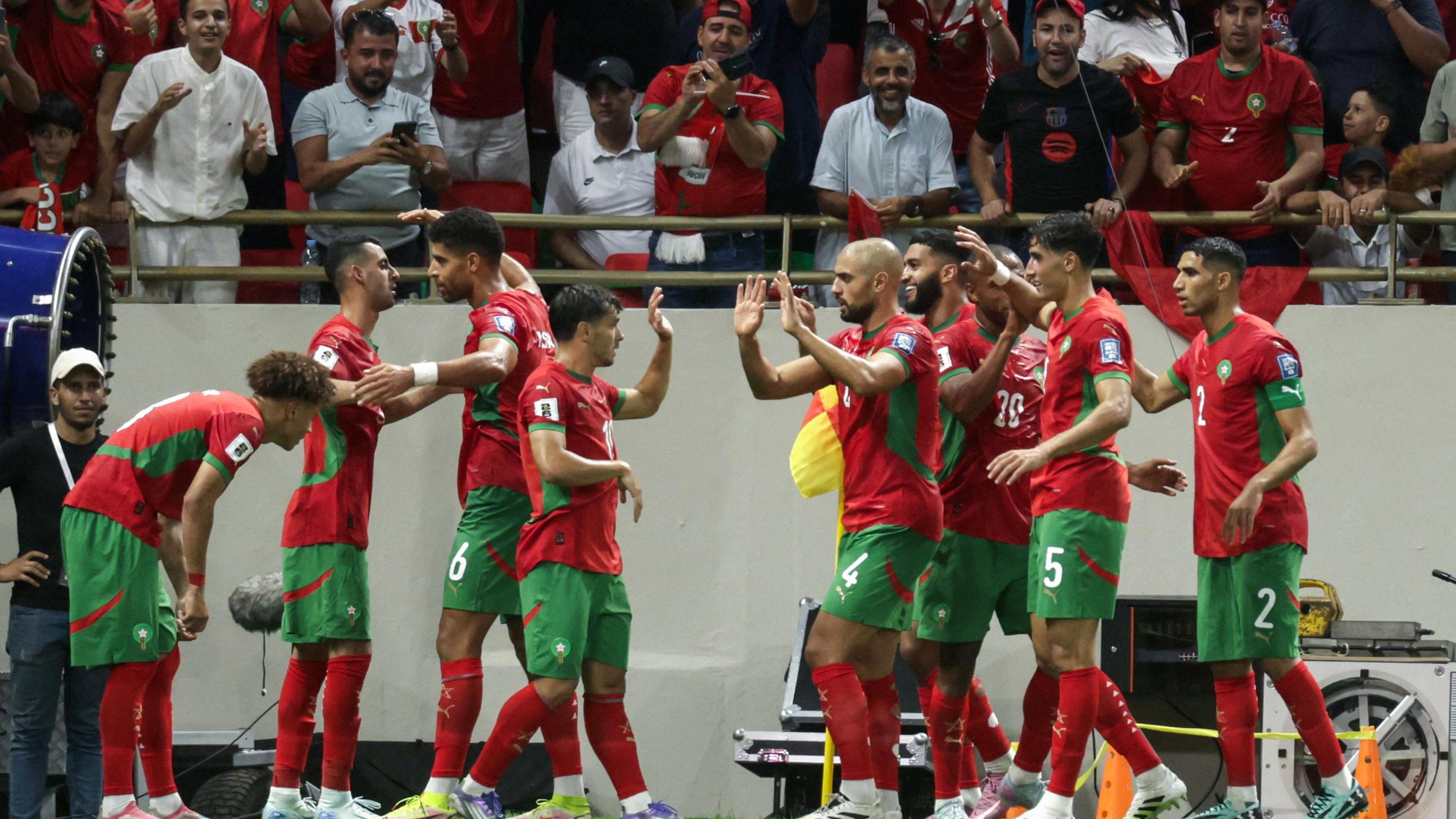 Nine Morocco players, all wearing a red and green strip, exchange high fives as they celebrate a goal, with the first two rows of Morocco fans seen cheering in the background