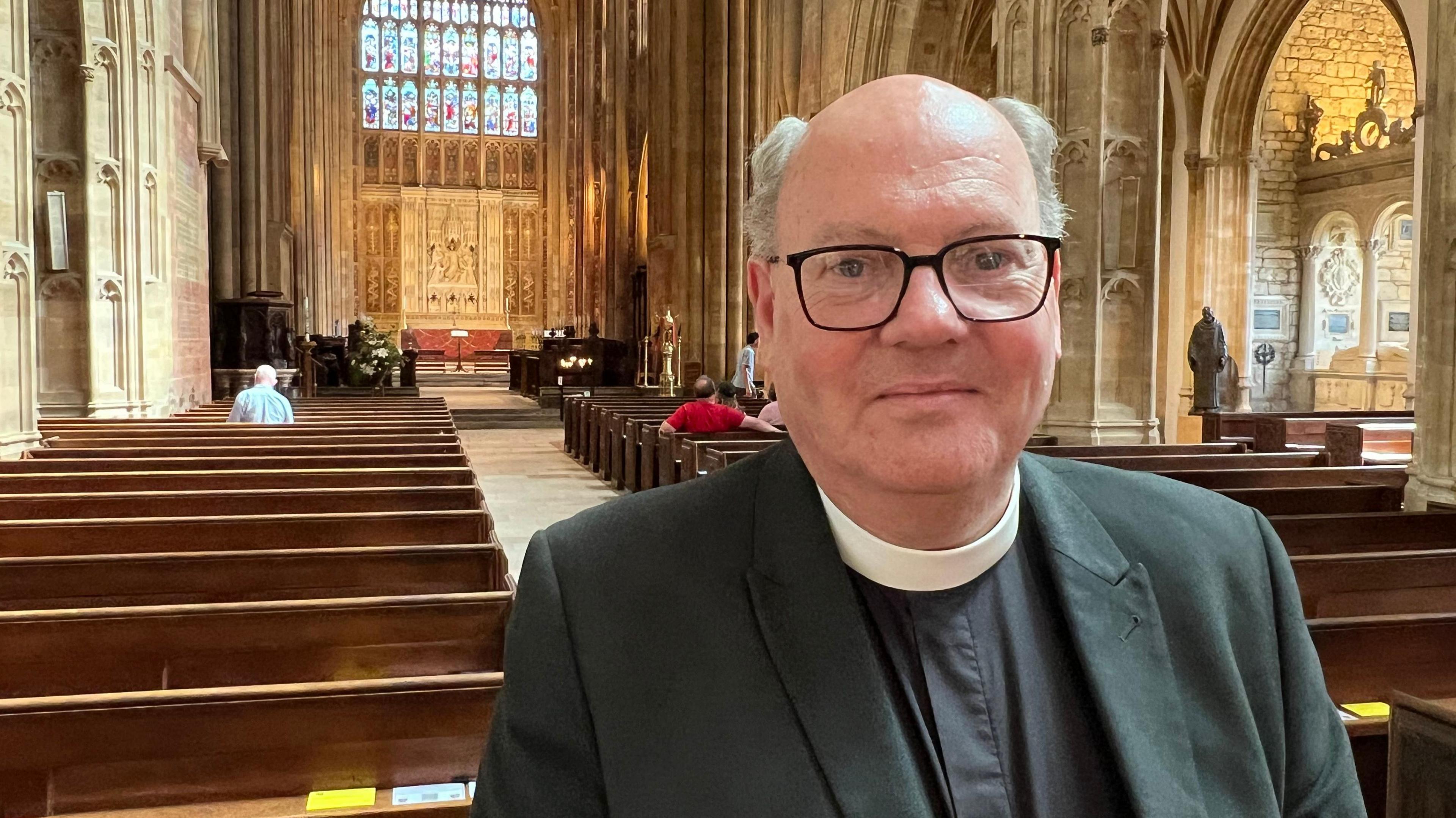 Rev Martin Lee wearing clerical collar in front of Sherborne Abbey pews and stained glass window in the background