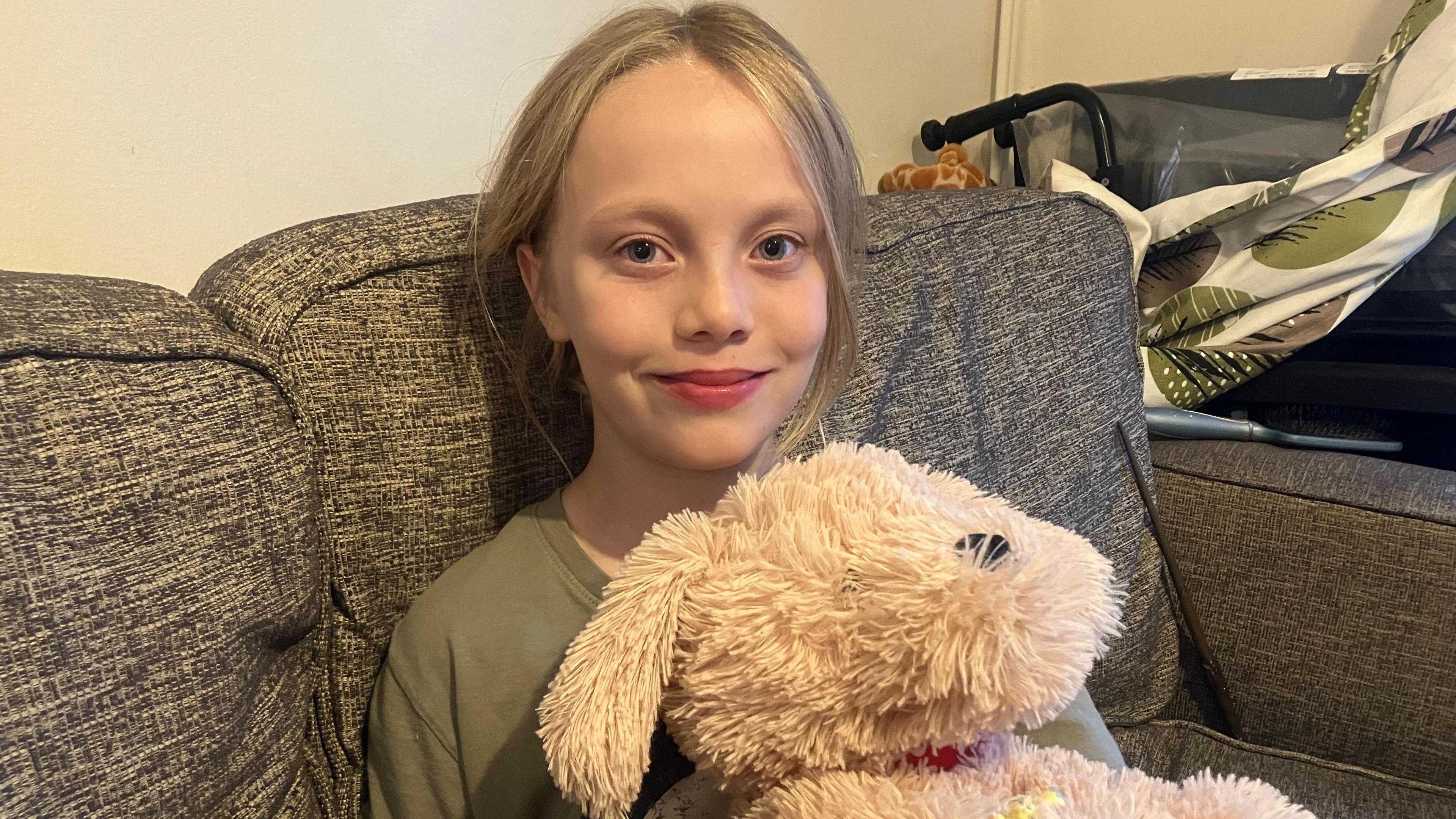 Aderyn is sitting on the sofa and smiling at the camera. She is wearing a khaki top with her blonde hair tied back. She is holding a dog teddy.