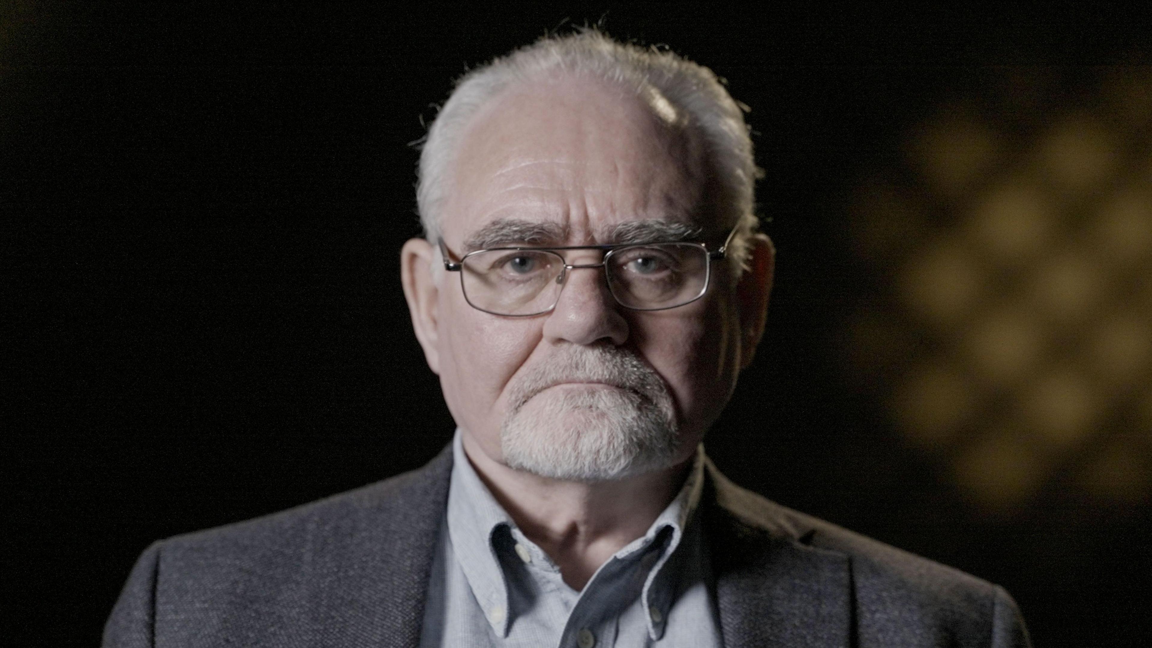 Patrick Magee, a man with a grey shirt, grey jacket, glasses and short grey hair and a short grey beard.