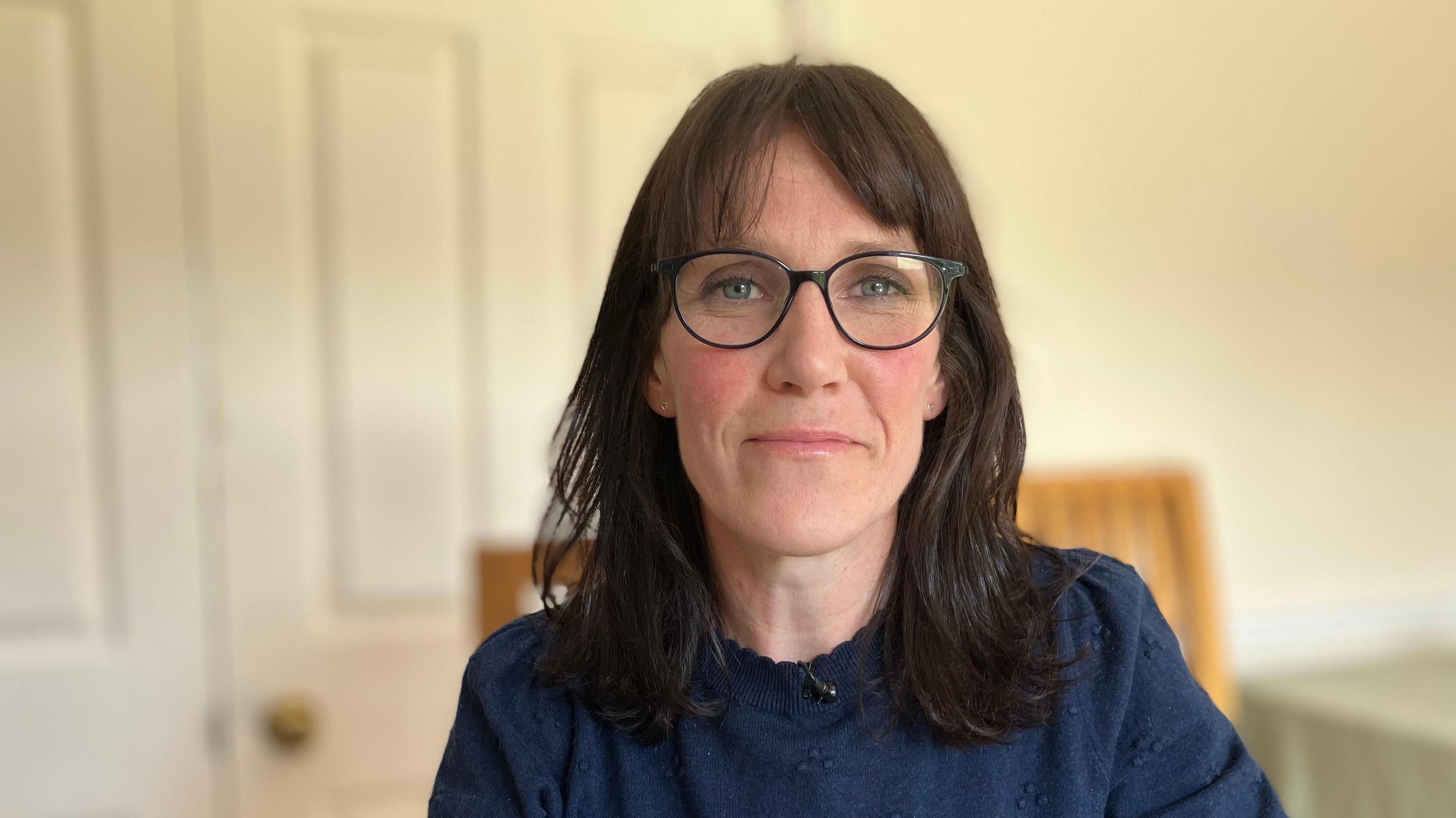 Dr Rachel Rutter is sat on a chair wearing a navy blue jumper. She has dark brown hair and is wearing black framed glasses and has a slight smile. 