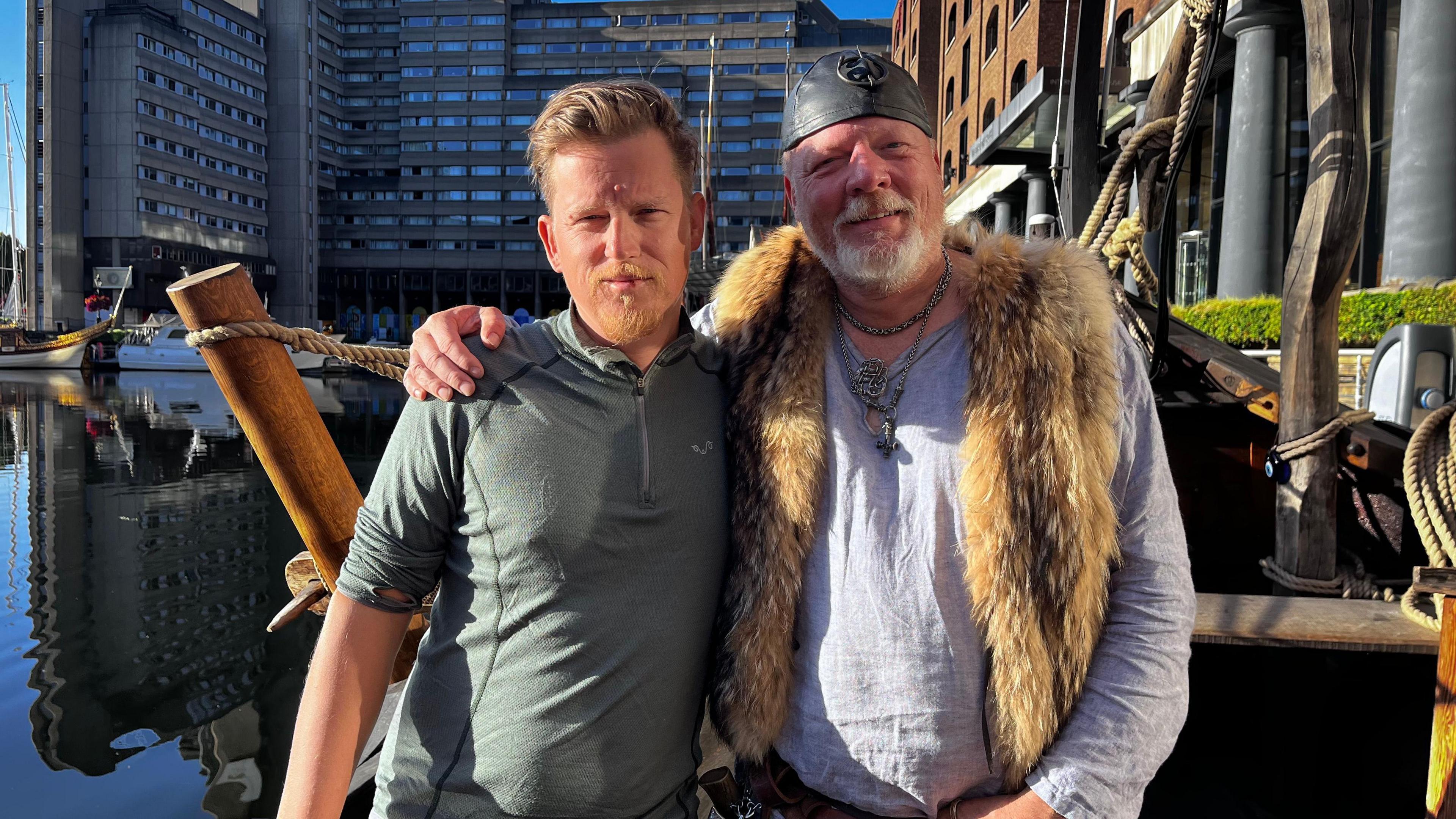 Two men are stood looking into the camera, with their arms around each other's shoulder, visible from the waist up. They are stood on a boat in dock, with a large grey building behind them. The man on the left has blonde hair and a short blonde beard, and is wearing a grey-green fitness top. The man on the right is smiling and wearing a Viking-style hat, and has a grey long-sleeve top and fluffy vest.