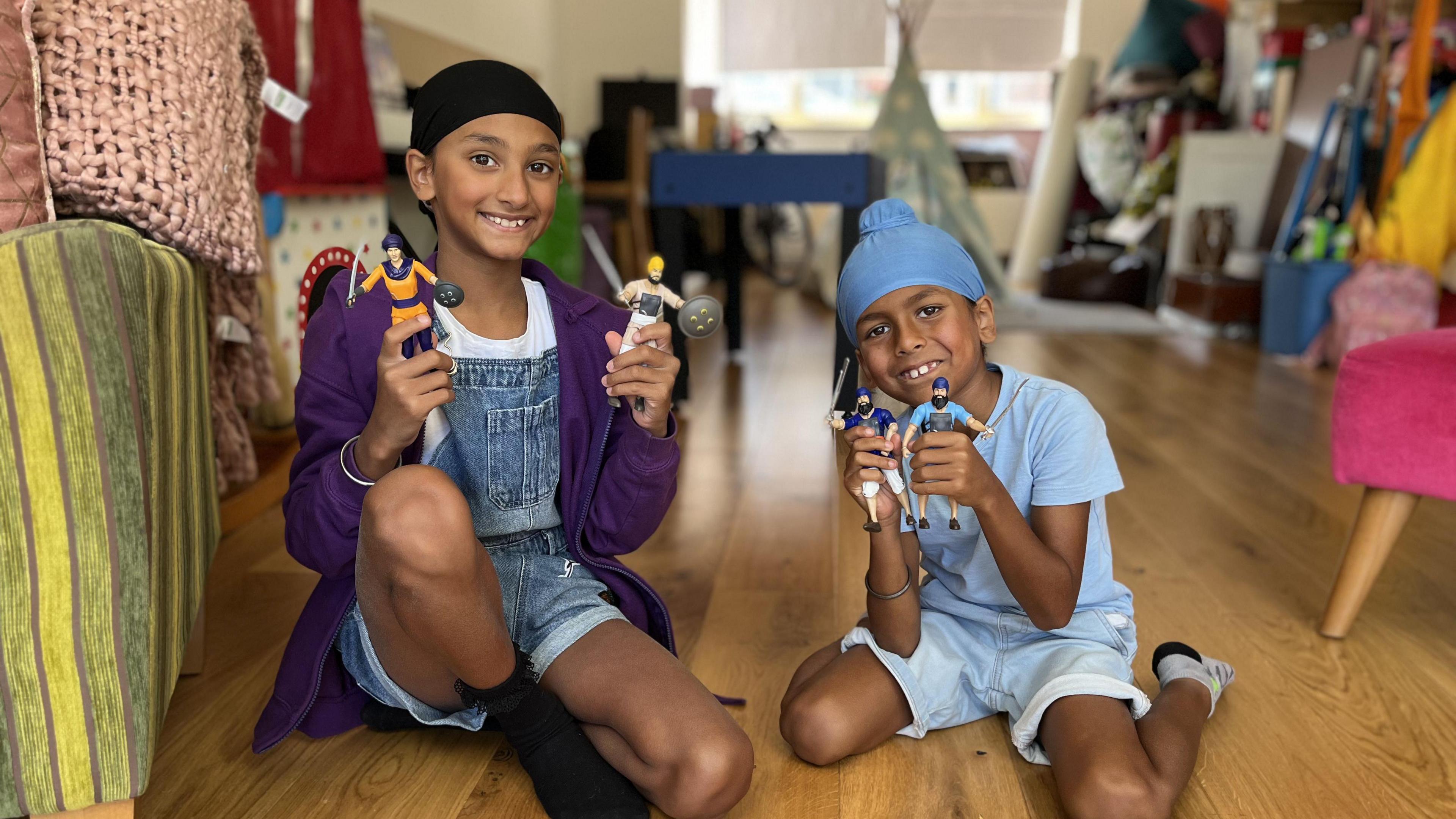 A girl and a boy holding an action figure in each hand, smiling at the camera in the room of a house