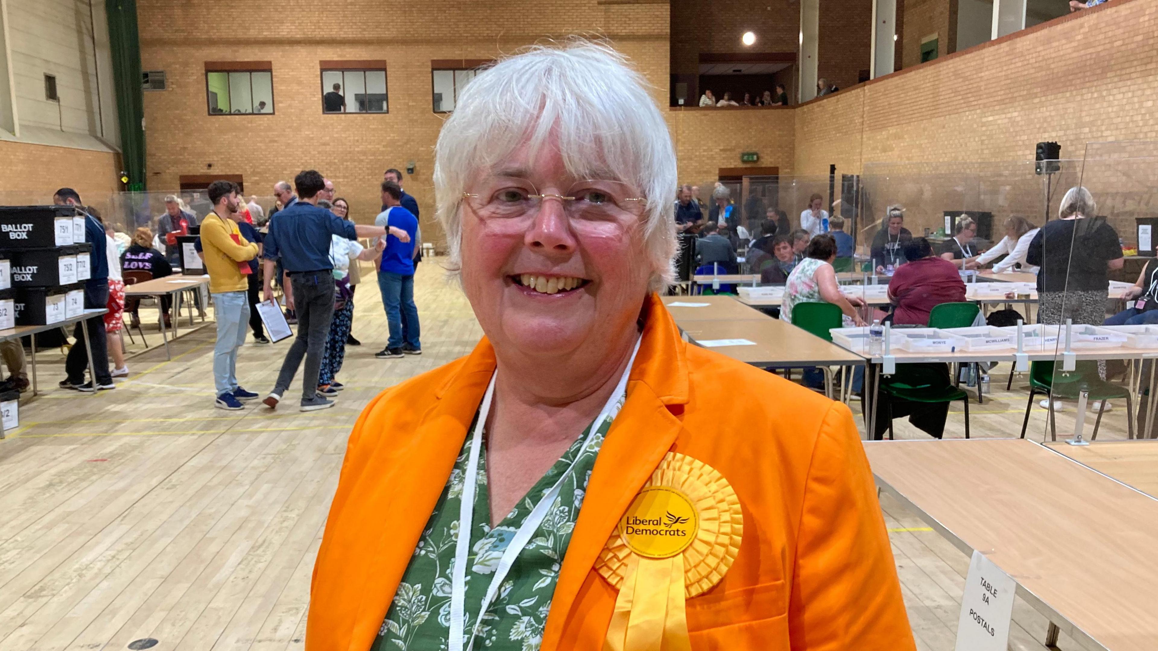 Picture of Charlotte Cane wearing an orange jacket, a liberal democrat rosette, a green dress and white lanyard. In the background are ballot boxes and people counting votes at tables. 