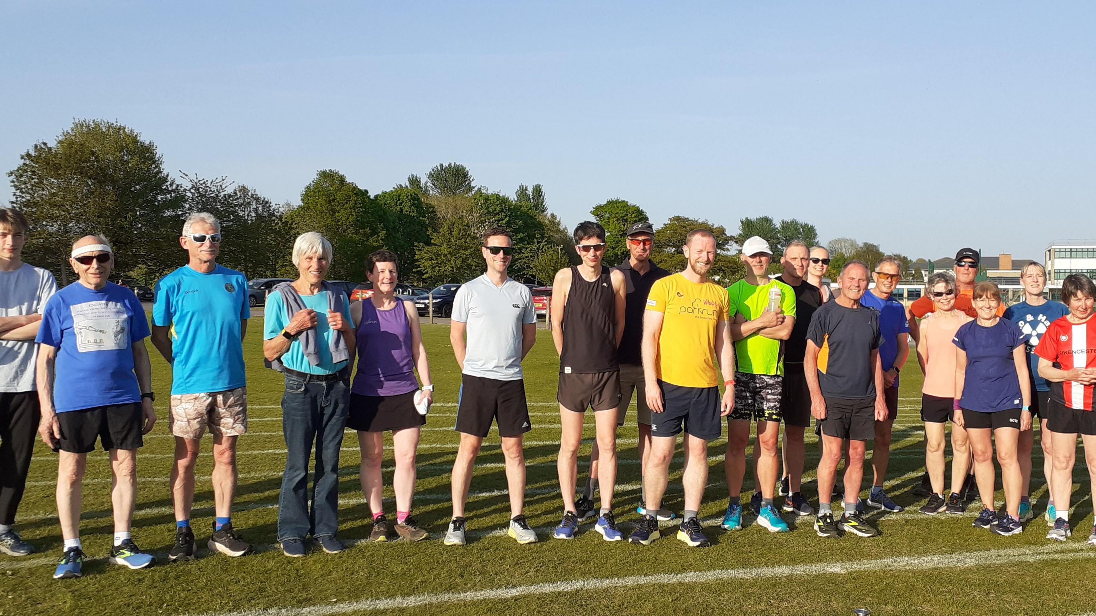 Runners fromm Cirencester Athletics Club preparing to go for a run. They are wearing a variety of shorts and t shirts and trainers. There are 19 male and female runners of varying adult ages and they are standing on an athletics track made of grass.