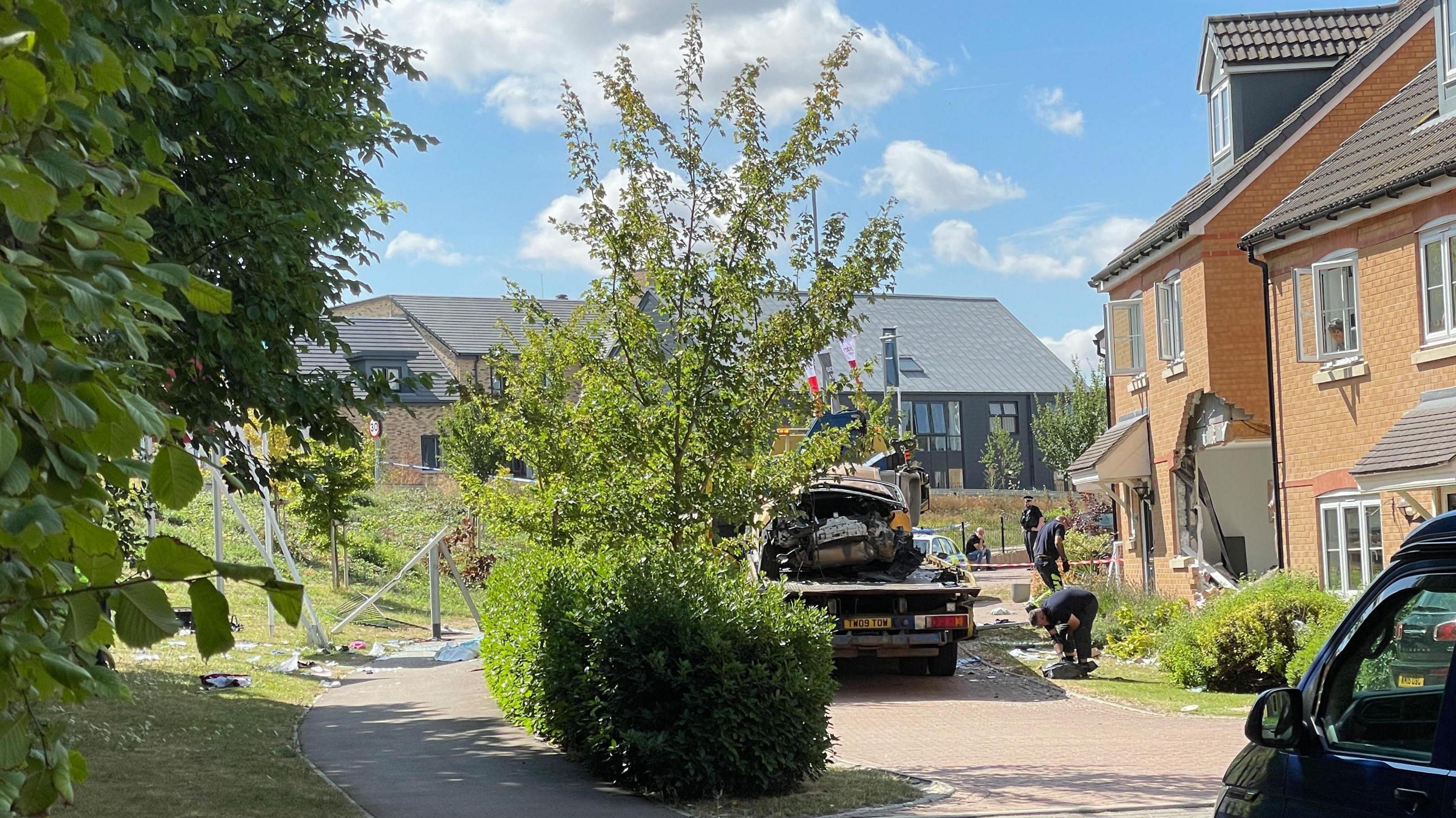 A house with its front wall collapsed. In front of it is a severely damaged car on the back of a recovery vehicle. There are men assessing the scene. There are more houses in the background with a small tree and a hedge in the foreground.