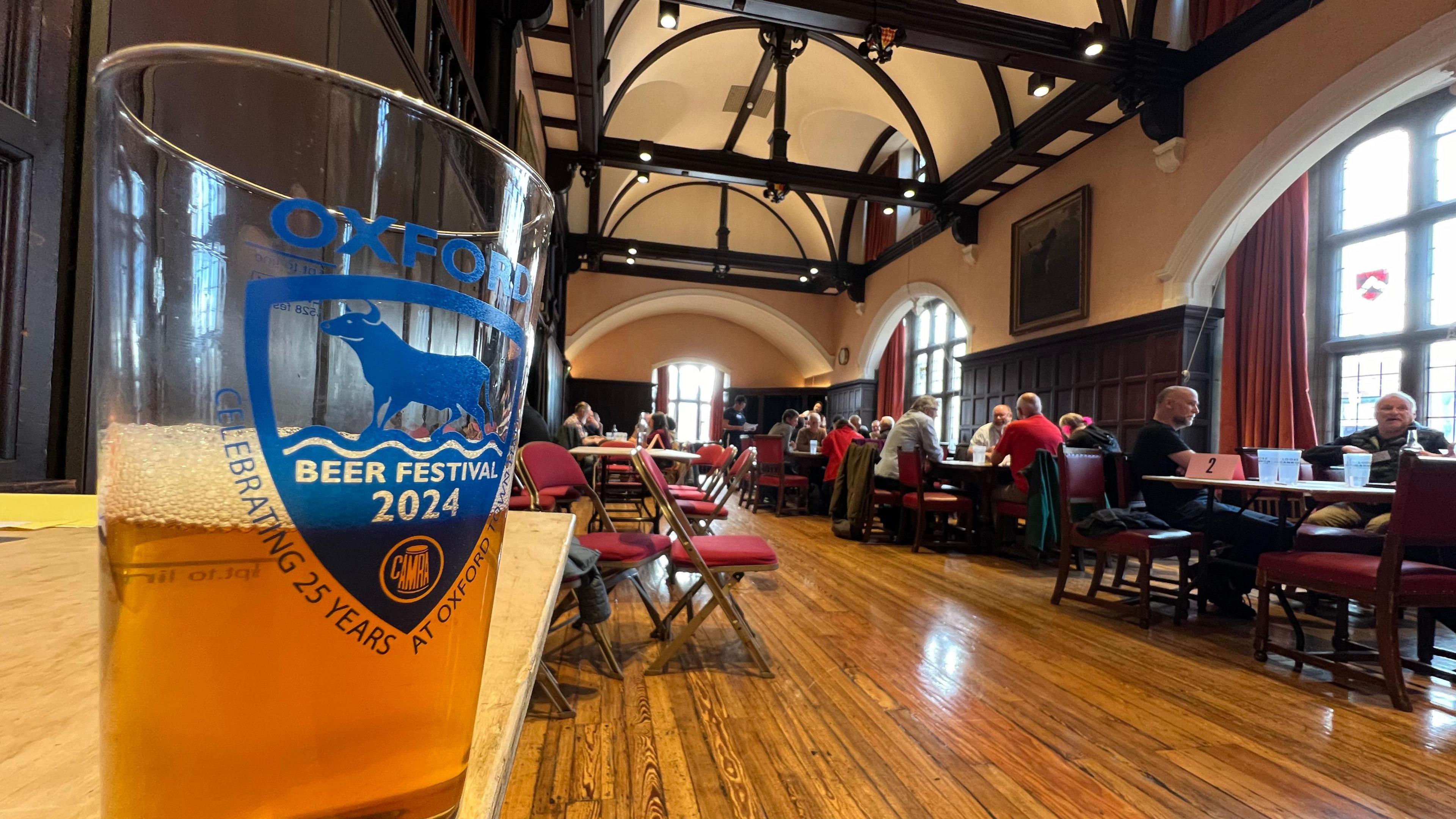 A view of a room at the town hall with people sitting on chairs at tables. There is a pint, half-full of beer, branded "Oxford Beer Festival 2024" on the glass. 