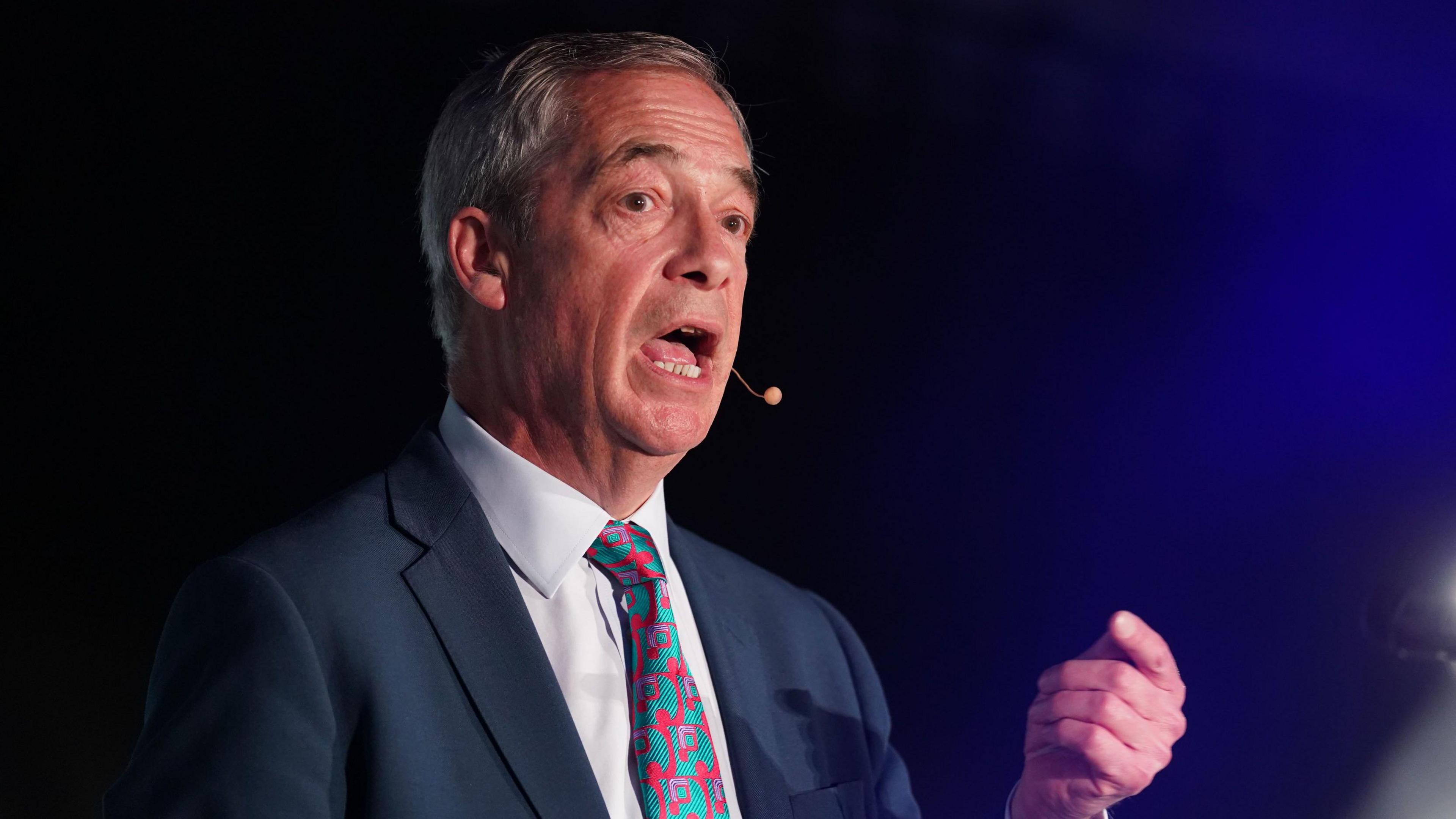Nigel Farage speaking while gesturing with his hand. He is wearing a blue suit and a red and green tie, with a white shirt.