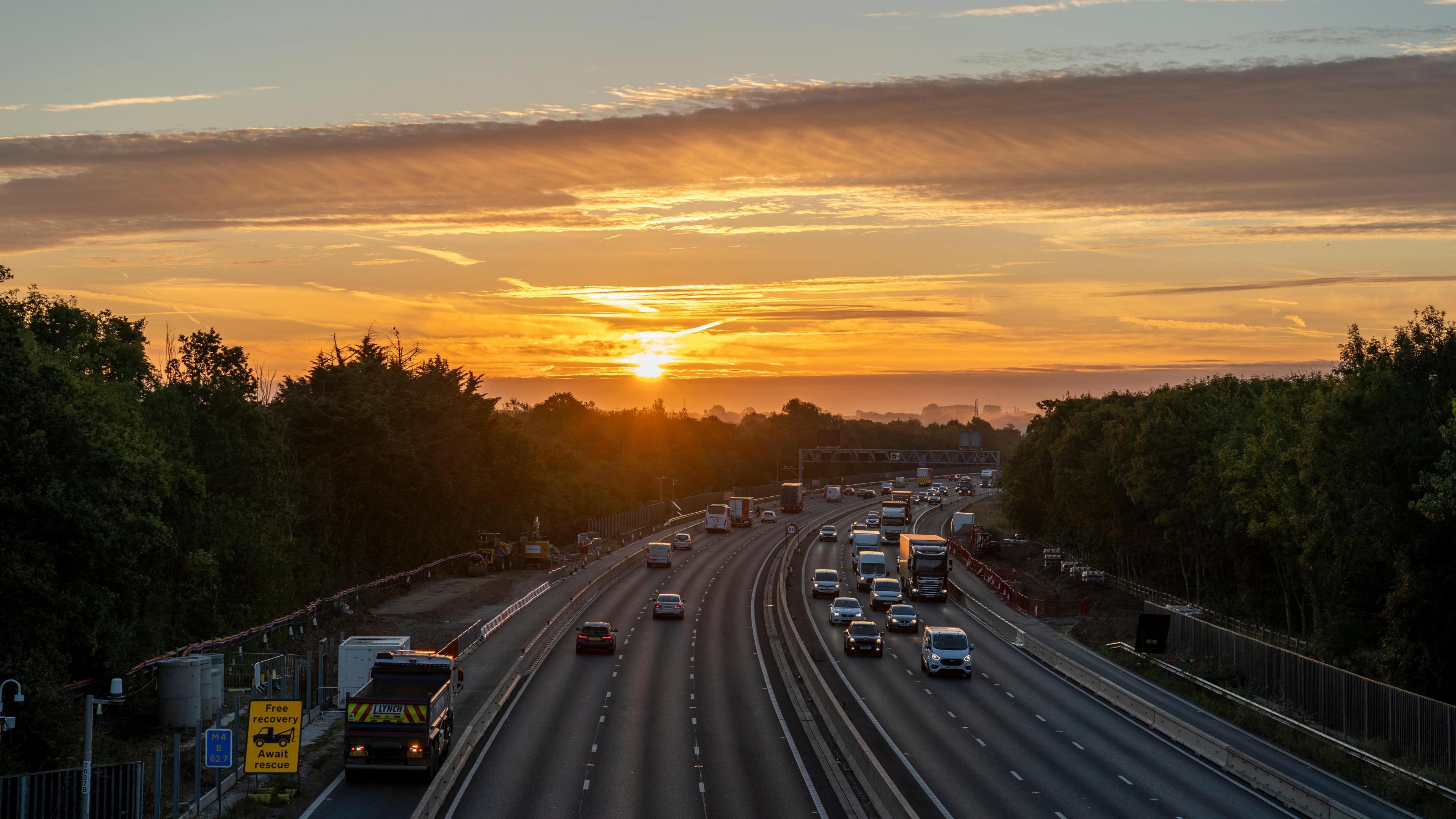 Two three-lane carriageways with several cars and lorries driving along are surrounded by a line of green trees on each side and overlooked by a low orange sun piercing through the clouds. 