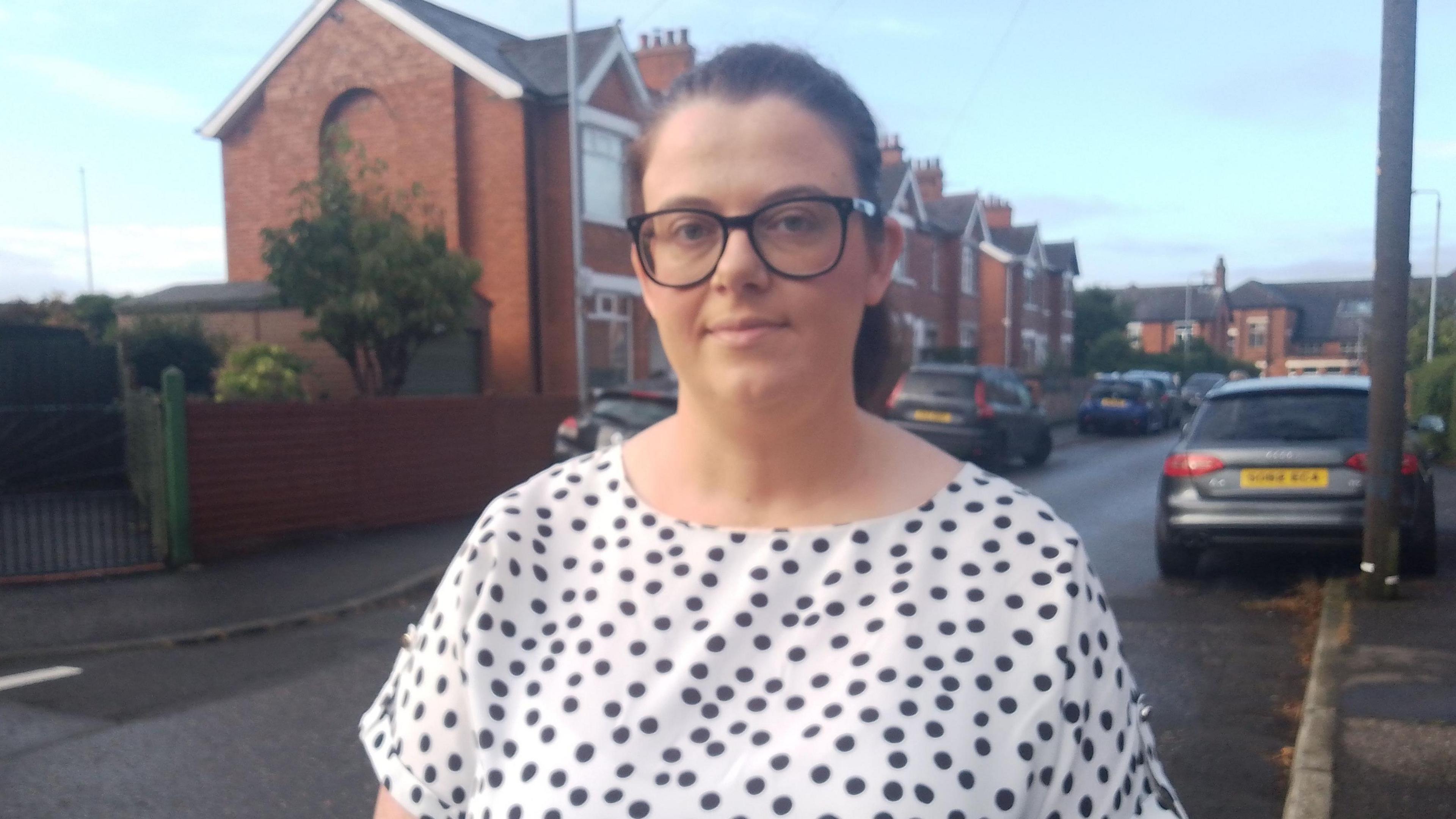 Ruth Brooks standing on a residential street, she has tied up brown hair, wearing black framed glasses and a white with black polka dotted t-shirt