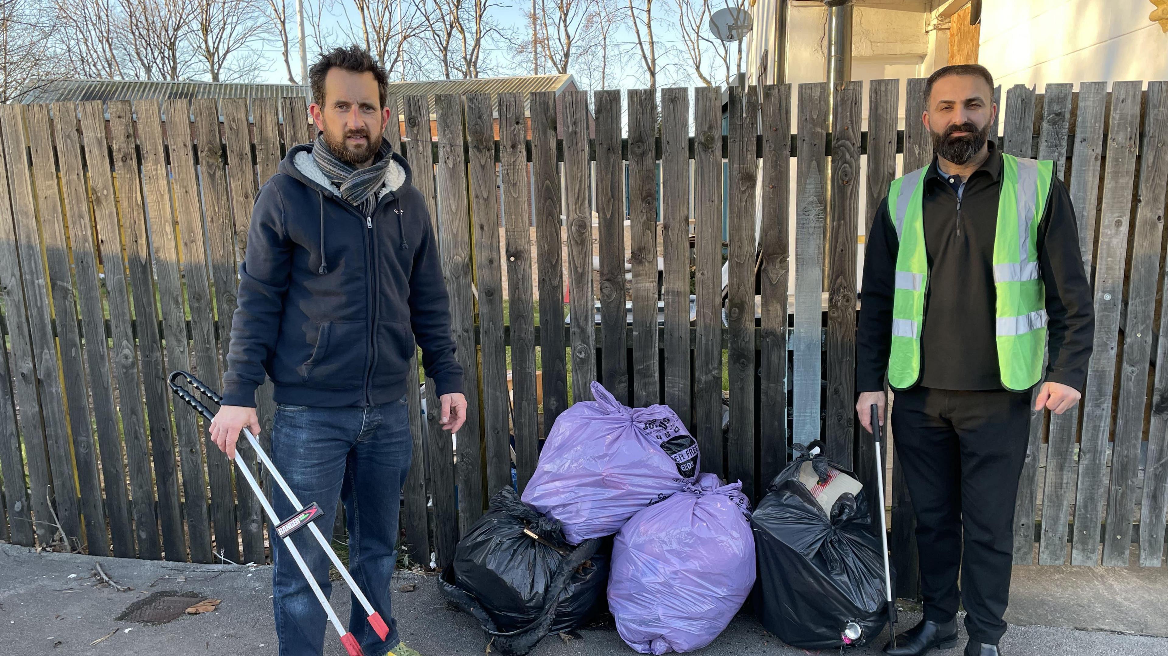 A man wearing a scarf and a dark hoodie and a man wearing a luminous green bib stand either side of some bags of rubbish. A wooden garden fence is in the background and they are both carrying litter picking tools.     