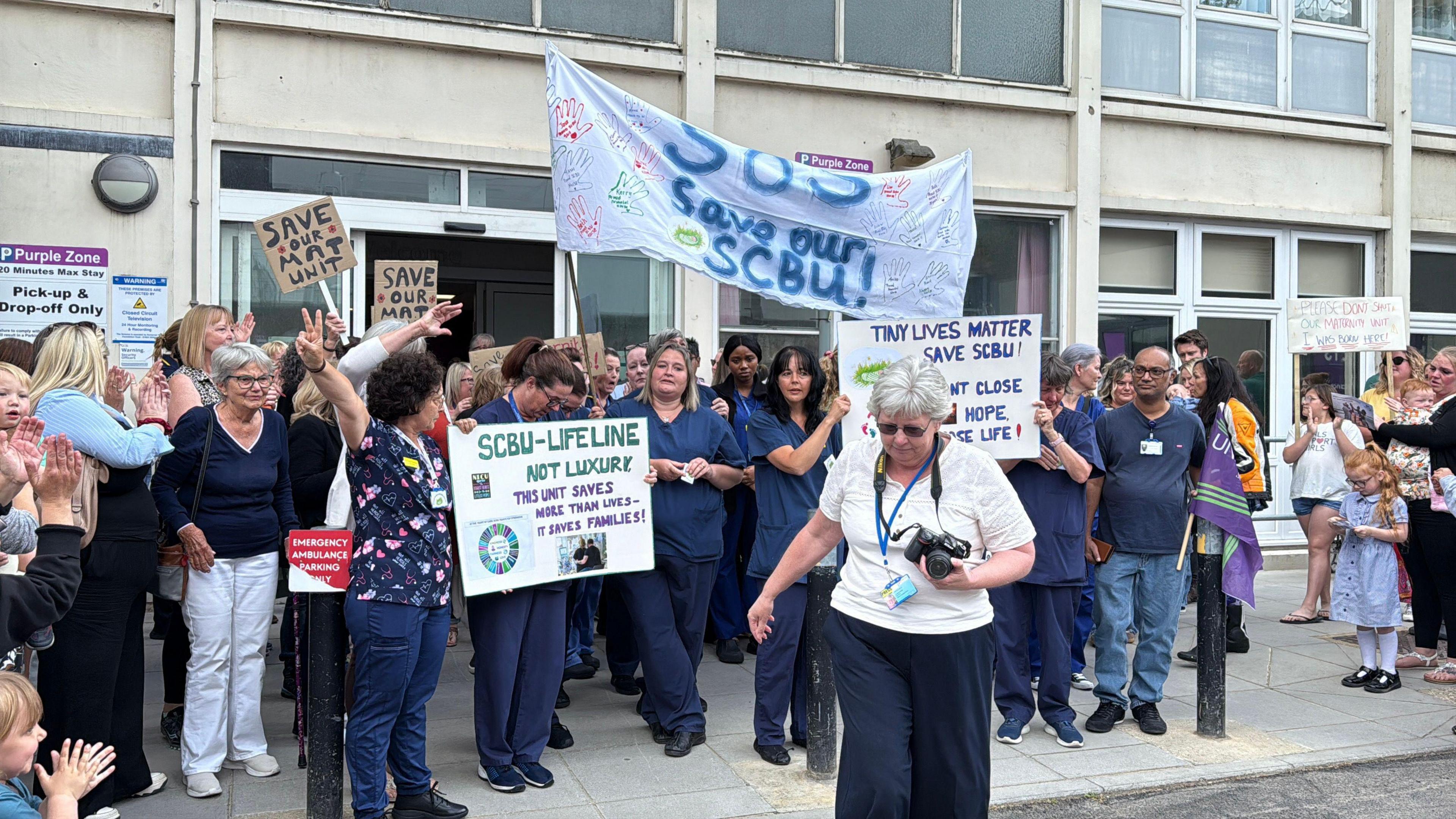 A group of people holding signs and banners, including 'save our mat unit' outside the exterior of the hospital.
