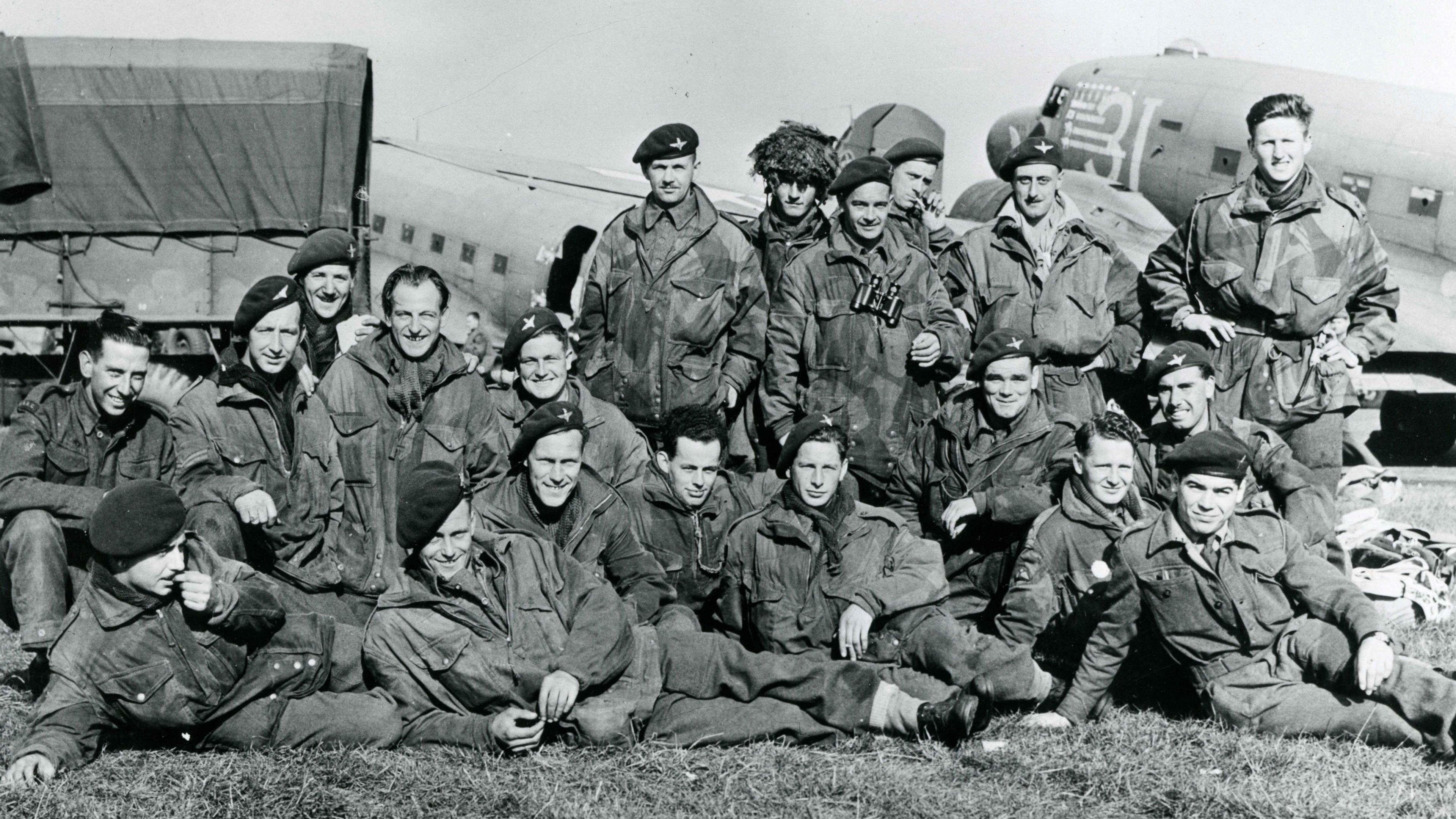 A black and white image of a group of soldiers sitting on the grass in uniform in front of a lorry and an aircraft.