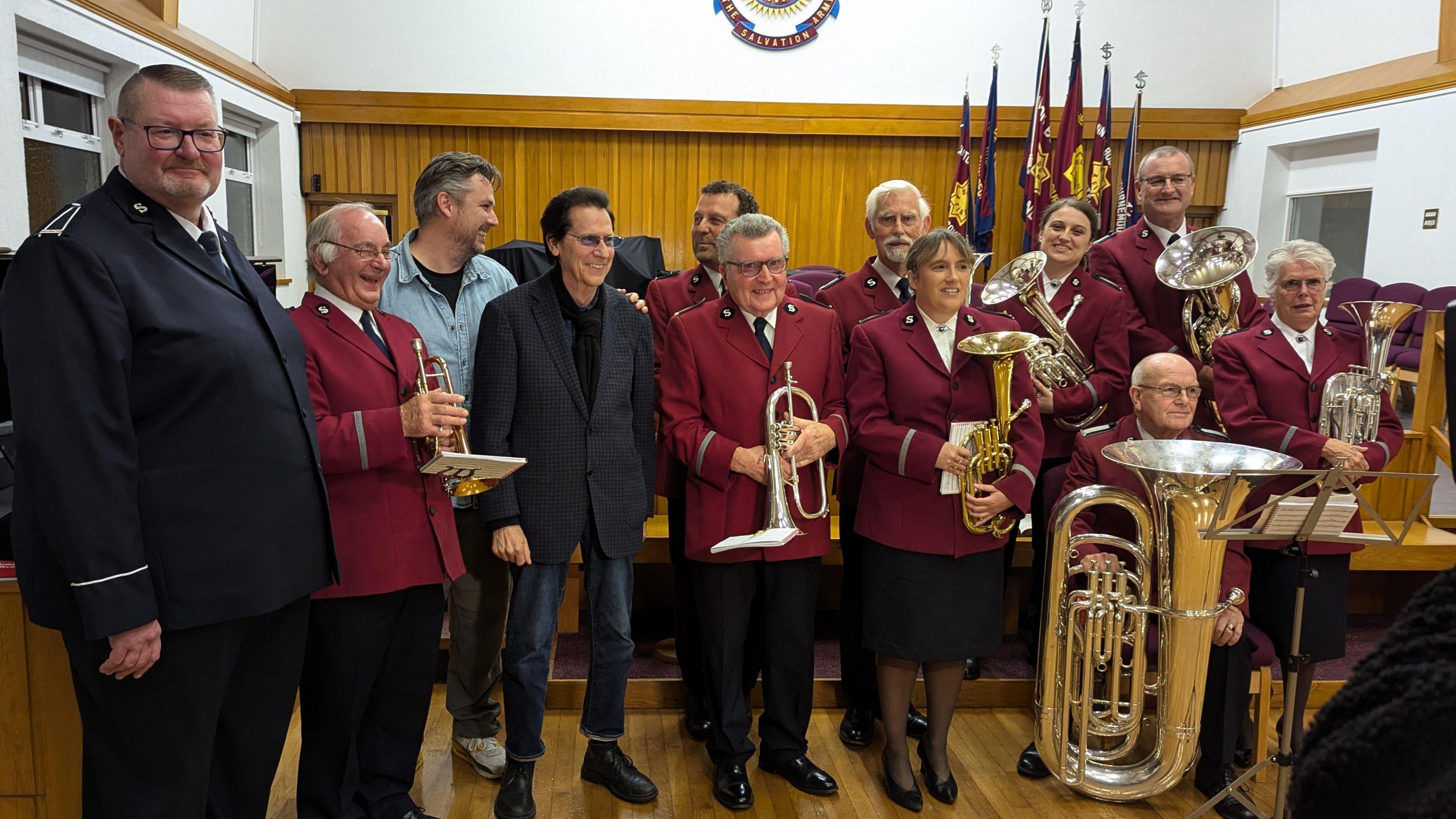 Shakin' Stevens stood in the centre wearing a blazer and some jeans next to people from the charity and the brass band who are wearing red uniforms.