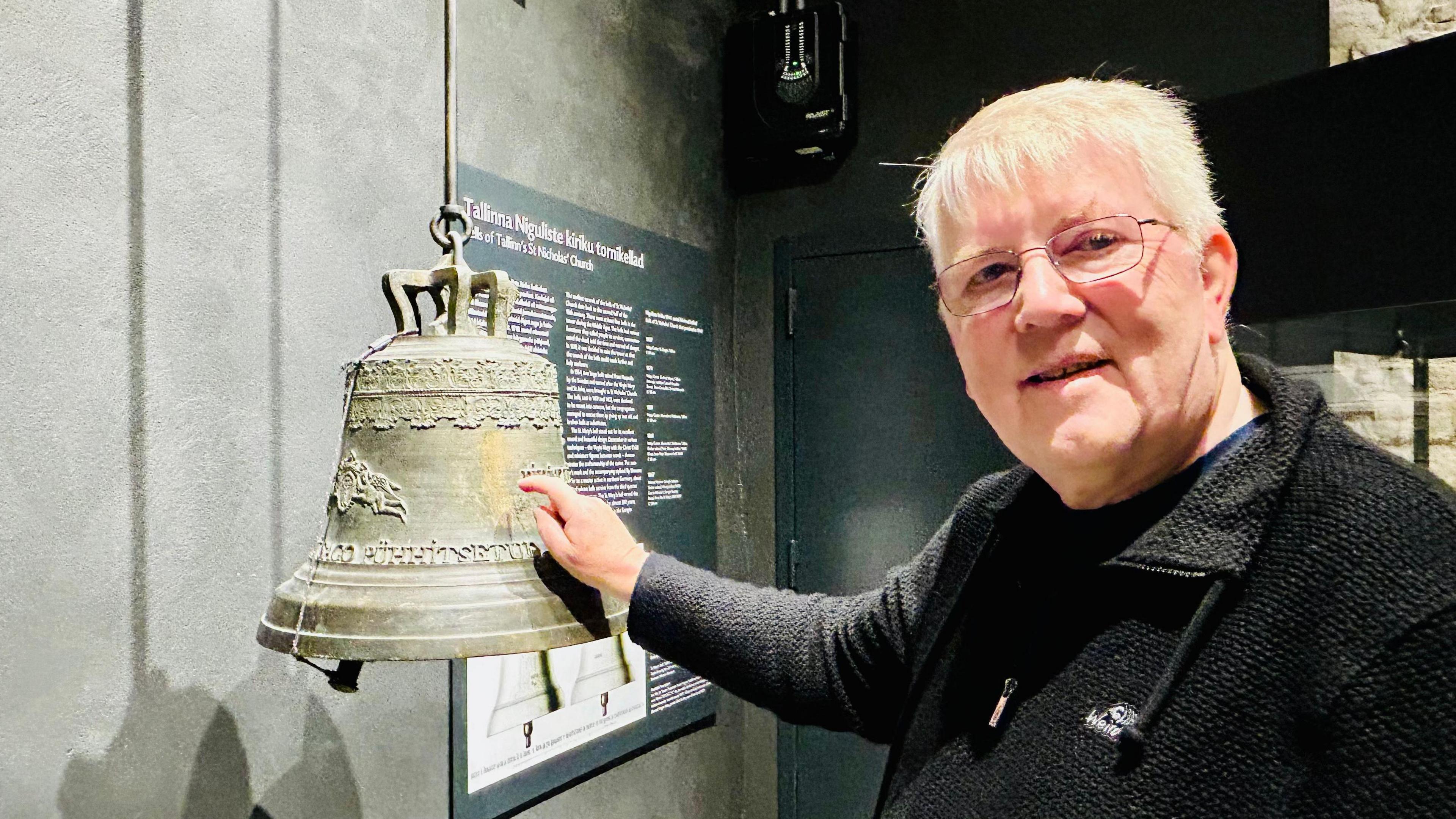 Chris Skinner is wearing glasses, has short grey hair and is wearing a black jacket over a dark jumper. His right hand is resting on a metal bell hanging against a grey wall