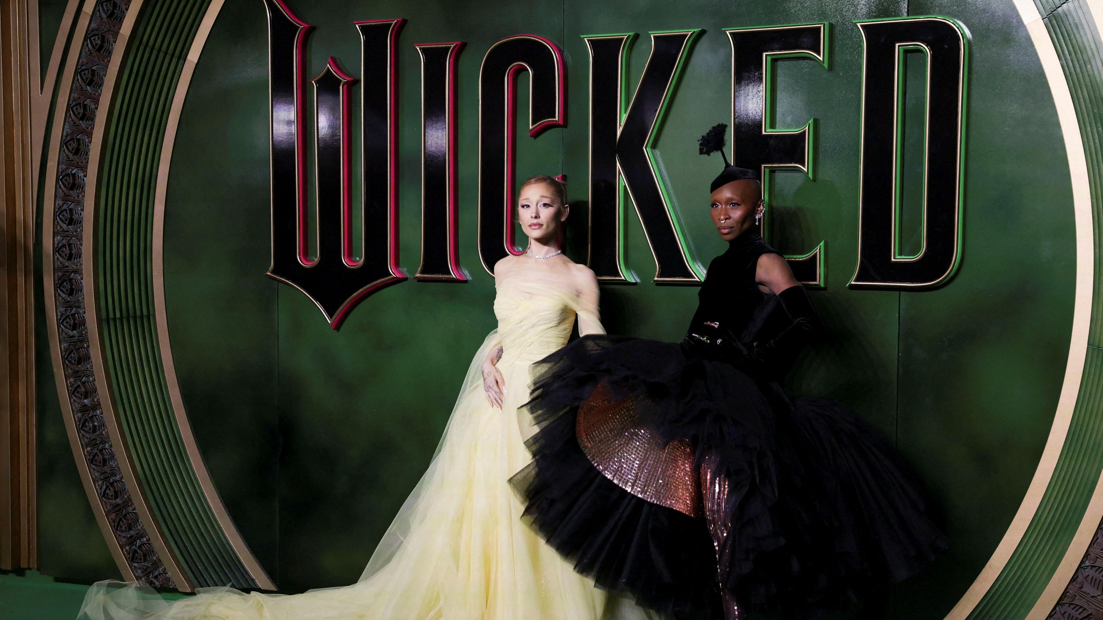 Ariana wears a pale yellow gown and is standing next to Cynthia who is wearing a black gown with a hooped skirt which is lifted slightly to reveal glittery leggings and lining underneath. THey are standing in front of a Wicked sign.