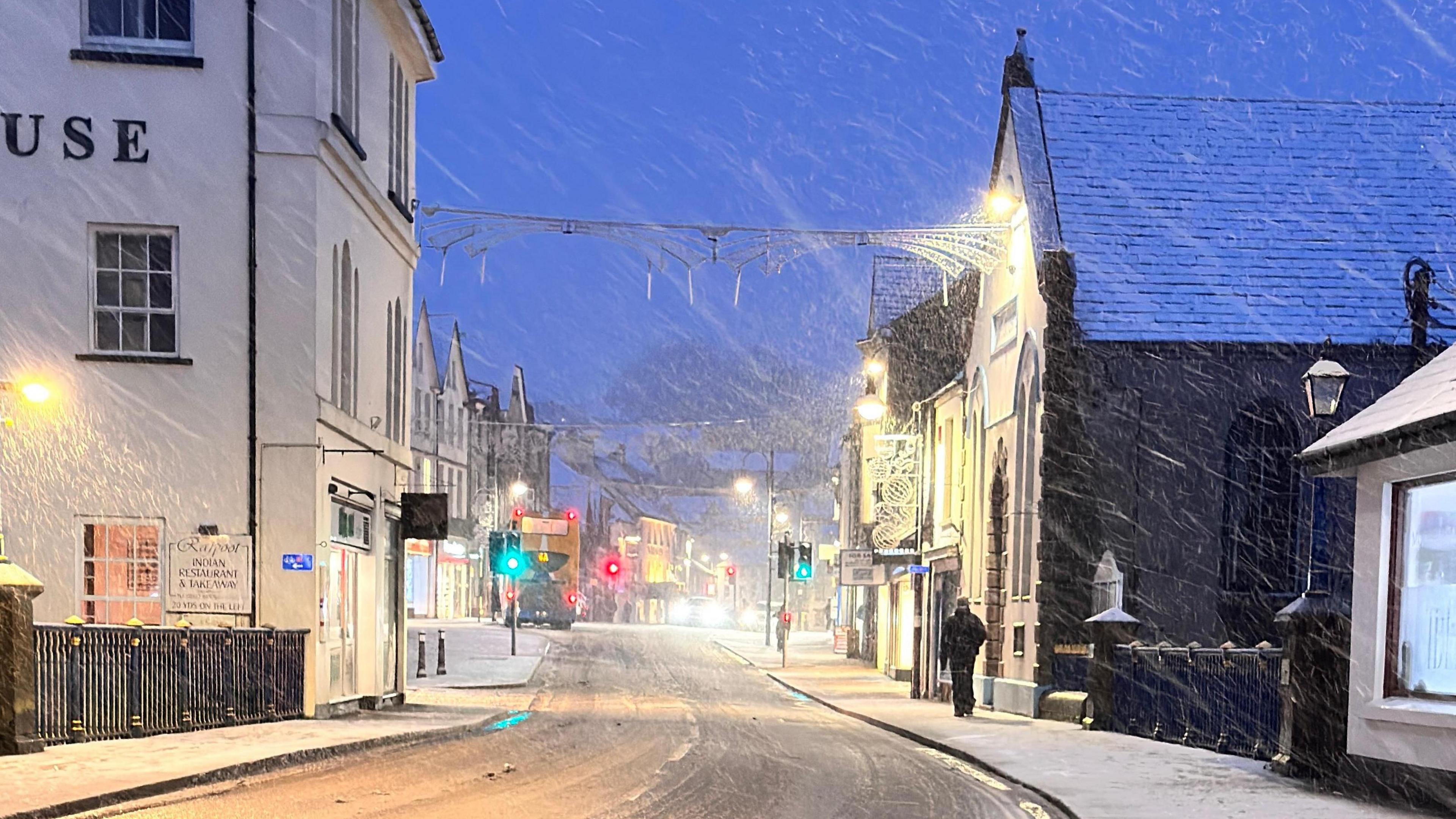 A high street with brick buildings on either side during snowfall in the early evening. The road and pavement are covered in snow.